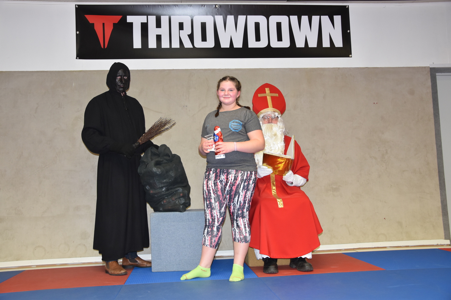 M&auml;dchen mit Wasserflasche steht zwischen einem schwarzen, maskierten Mann und einem rot gekleideten Nikolaus.