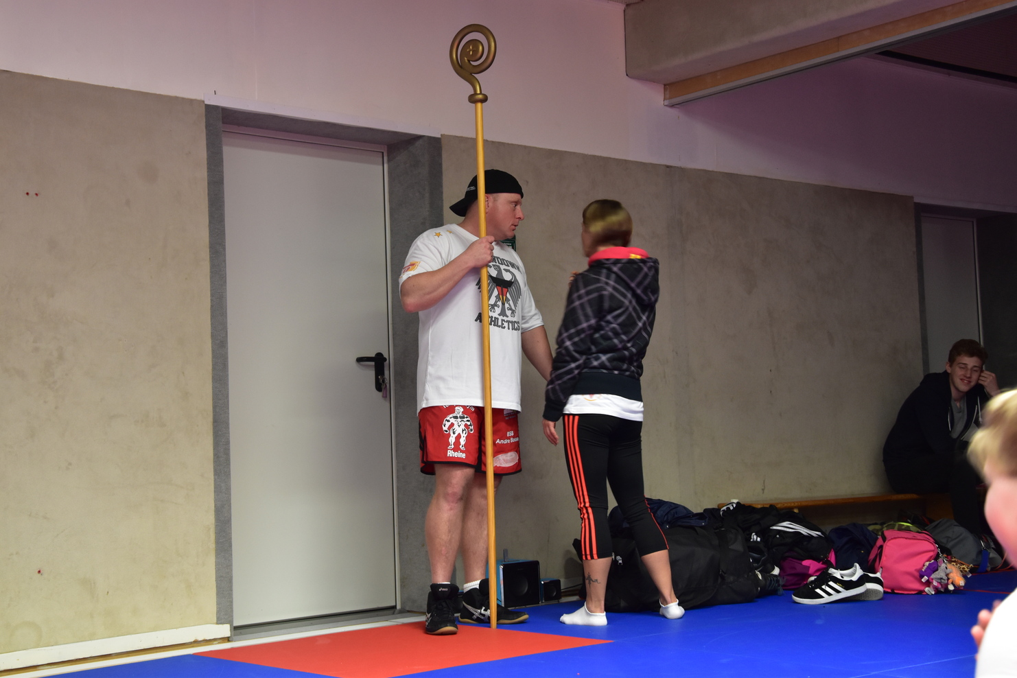 Trainer im T-Shirt und Shorts spricht mit einem M&auml;dchen, w&auml;hrend er einen Trainingsstab h&auml;lt, in einer Sporthalle.