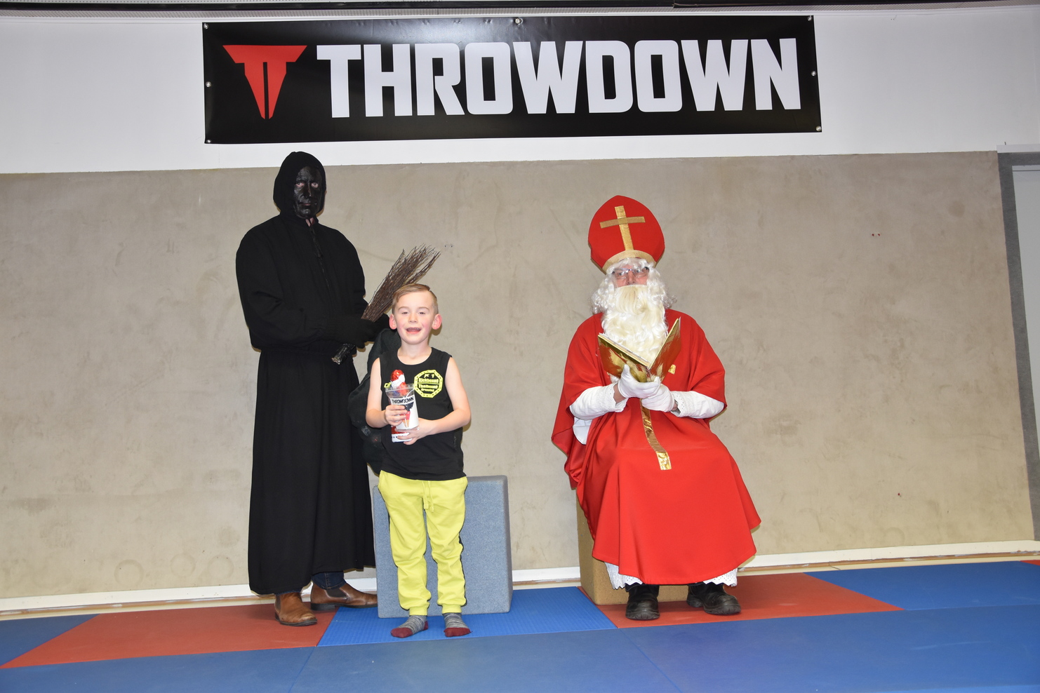 Junge mit Pokal steht zwischen einer dunkel gekleideten Figur und dem Nikolaus auf einer Sportmatte.