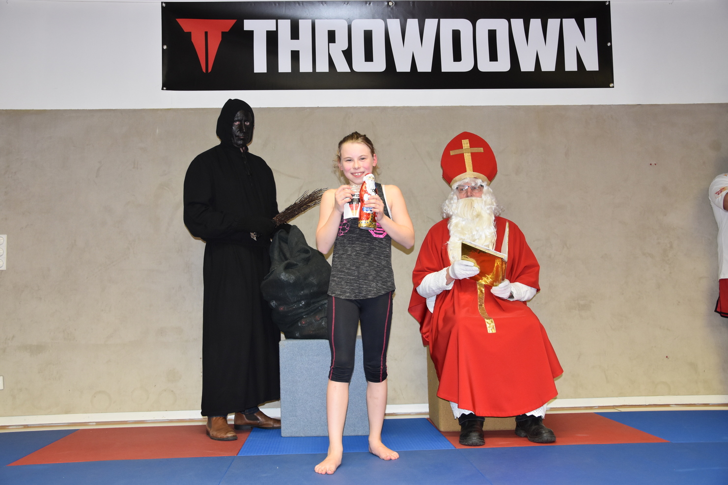 M&auml;dchen in Sportkleidung h&auml;lt eine Flasche, w&auml;hrend ein Kost&uuml;mierter und ein Mann in einem roten Gewand im Hintergrund stehen.