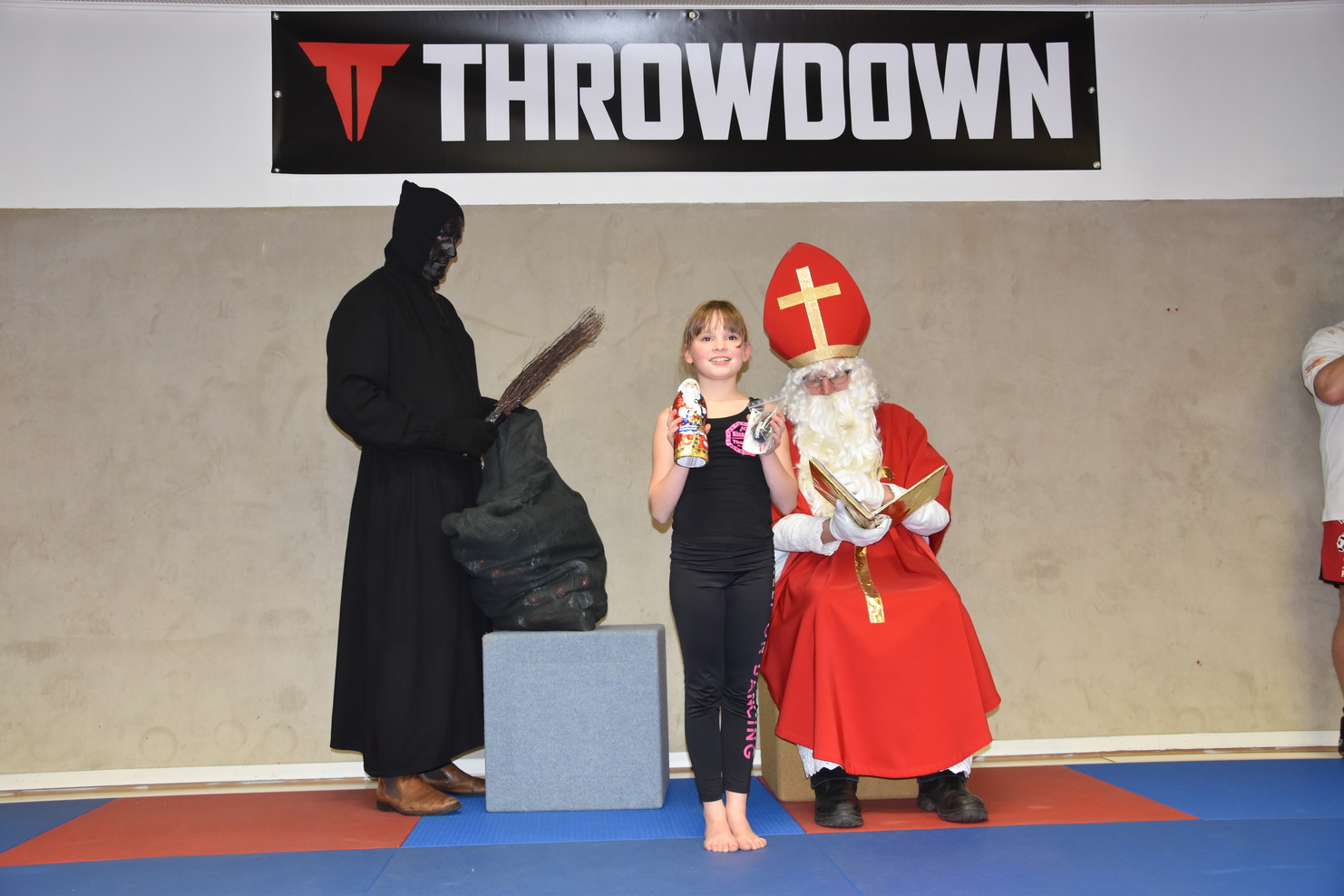 M&auml;dchen mit Getr&auml;nk steht zwischen einem schaurigen Wesen und einem Nikolaus auf einer Matte in einer Sporthalle.