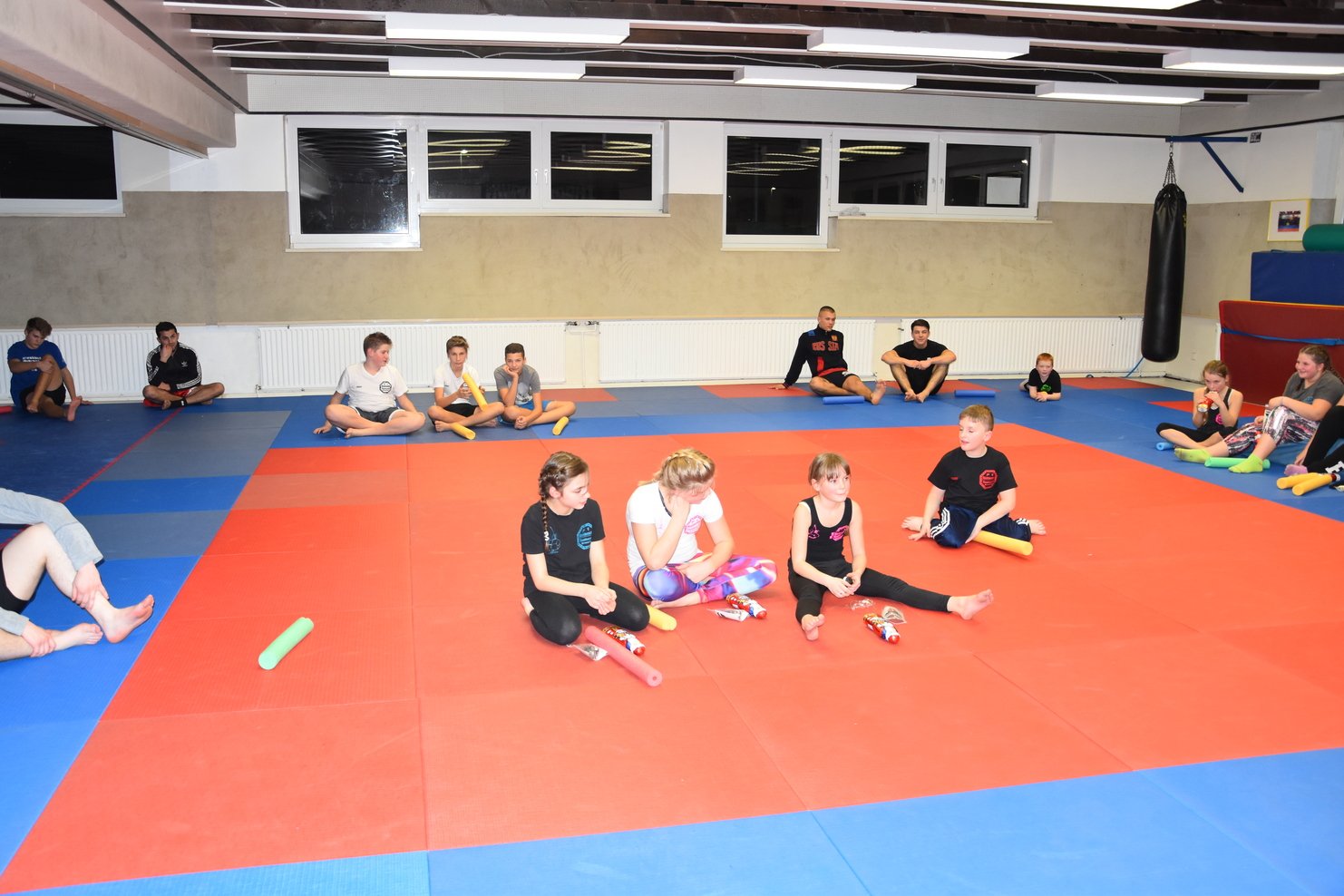 Gruppensitzung von Jugendlichen auf einer farbigen Trainingsmatte in einem Sportraum, einige mit Trainingsmaterial.
