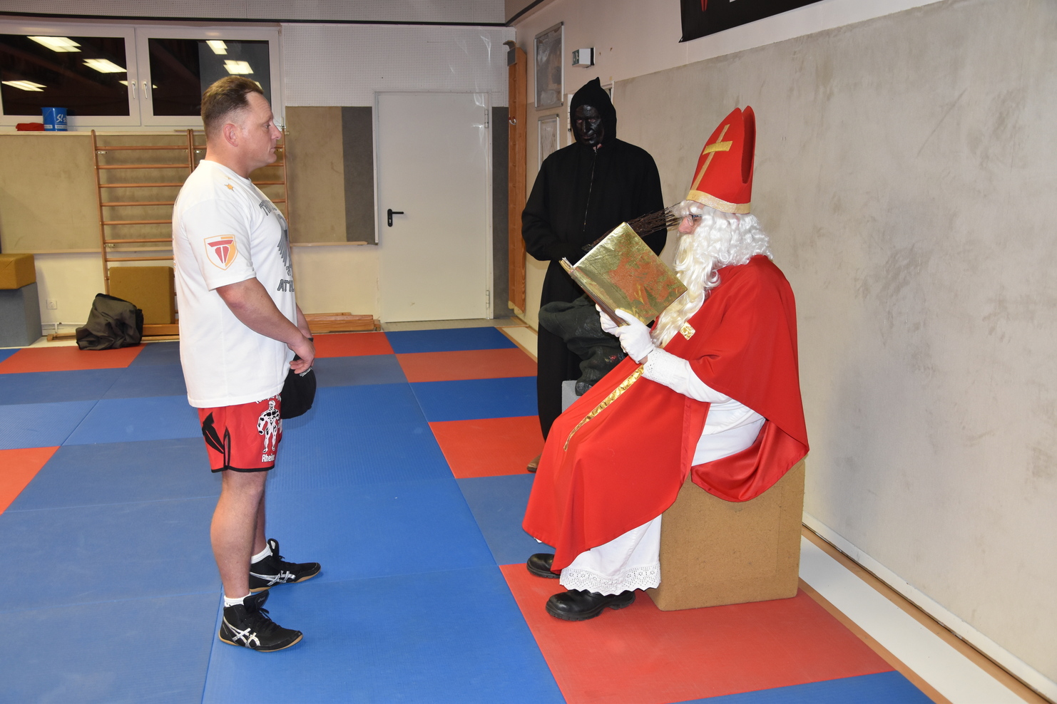 Eine Person im Sportoutfit steht vor einem sitzenden, verkleideten Nikolaus, w&auml;hrend eine weitere Person im Hintergrund zusieht.