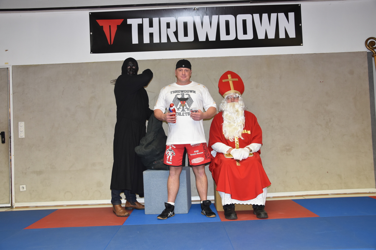 Drei Personen stehen auf einer blauen Matte: ein Mann in Sportkleidung, ein Heilige Nachtmann und eine maskierte Figur.