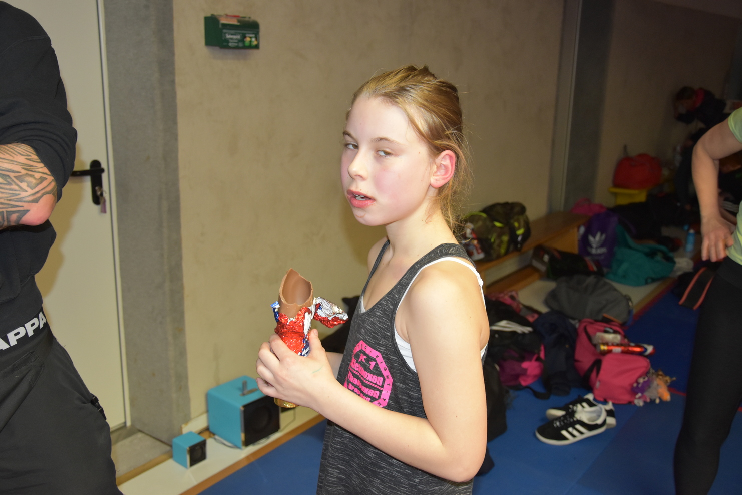 M&auml;dchen mit einem Schokoladenriegel in der Hand, steht in einem Sportraum mit Taschen und Schuhen im Hintergrund.