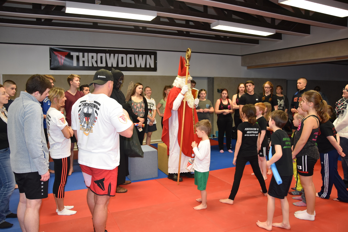 Eine Gruppe von Kindern und Erwachsenen steht um einen verkleideten Nikolaus in einem Trainingsraum mit Matten.