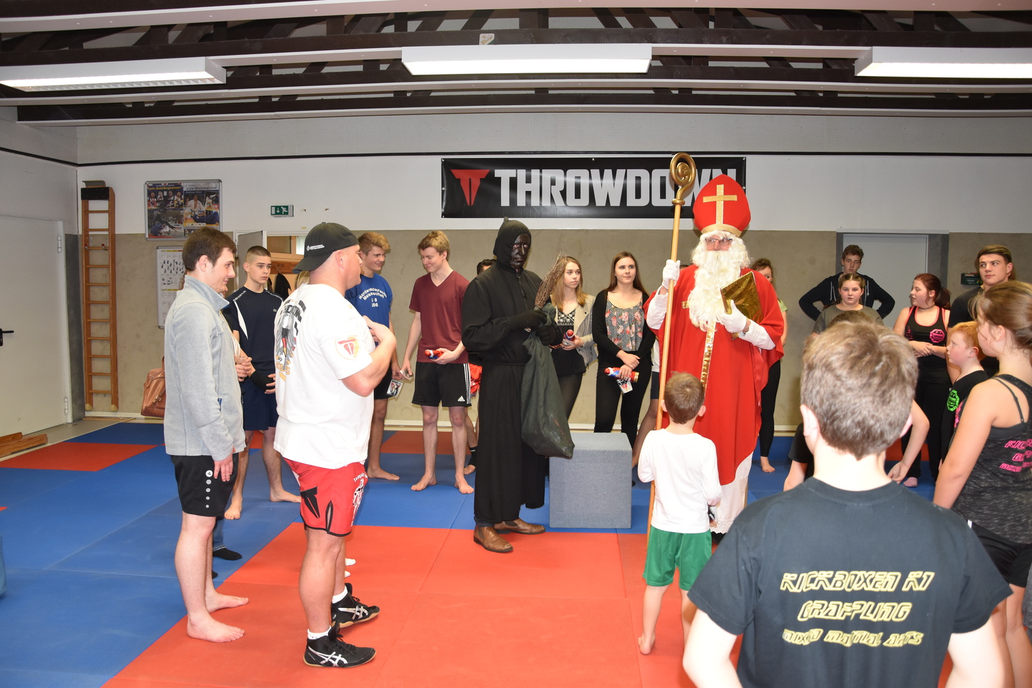 Gruppenszene in einem Trainingsraum: Nikolaus und Knecht Ruprecht begr&uuml;&szlig;en junge Sportler auf der Matte.