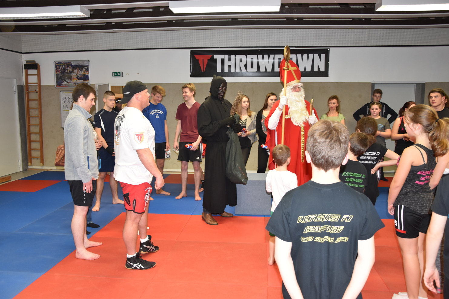 Gruppe von Personen in Sportkleidung steht um den Weihnachtsmann und einen verkleideten Helfer in einem Sportstudio.