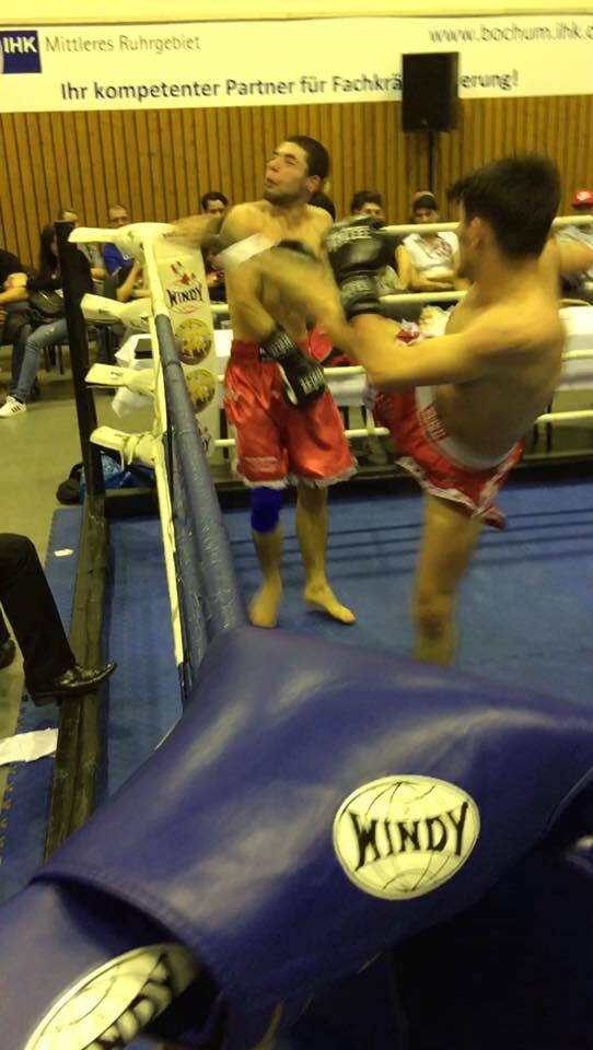 Zwei K&auml;mpfer in roter Shorts im Ring; einer wird mit einem Kick getroffen, Zuschauer im Hintergrund.