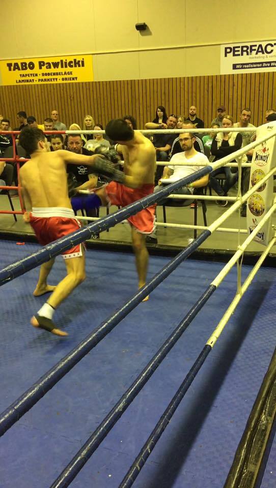 Zwei K&auml;mpfer im Boxring f&uuml;hren einen Kickboxkampf, w&auml;hrend Zuschauer am Rand zuschauen.