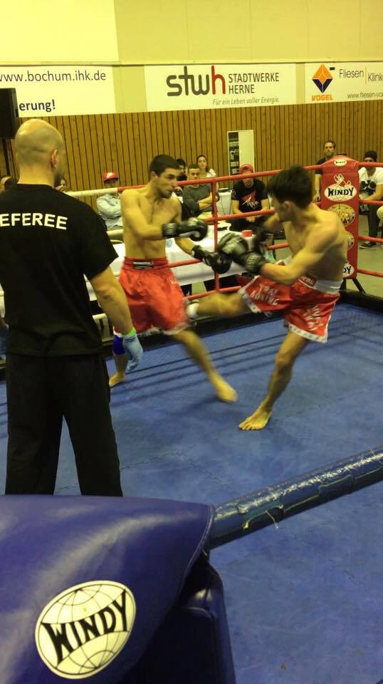 Zwei K&auml;mpfer im Boxring, einer tritt aus der Hocke, w&auml;hrend der Schiedsrichter zusieht. Zuschauer im Hintergrund.
