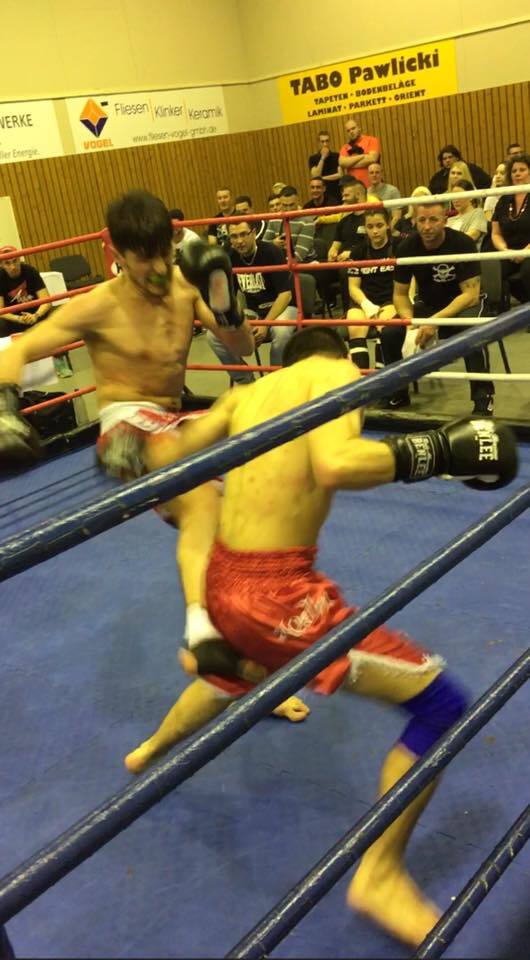 Zwei K&auml;mpfer im Ring, einer tritt w&auml;hrend eines Wettkampfs, Zuschauer verfolgen das Geschehen gespannt.