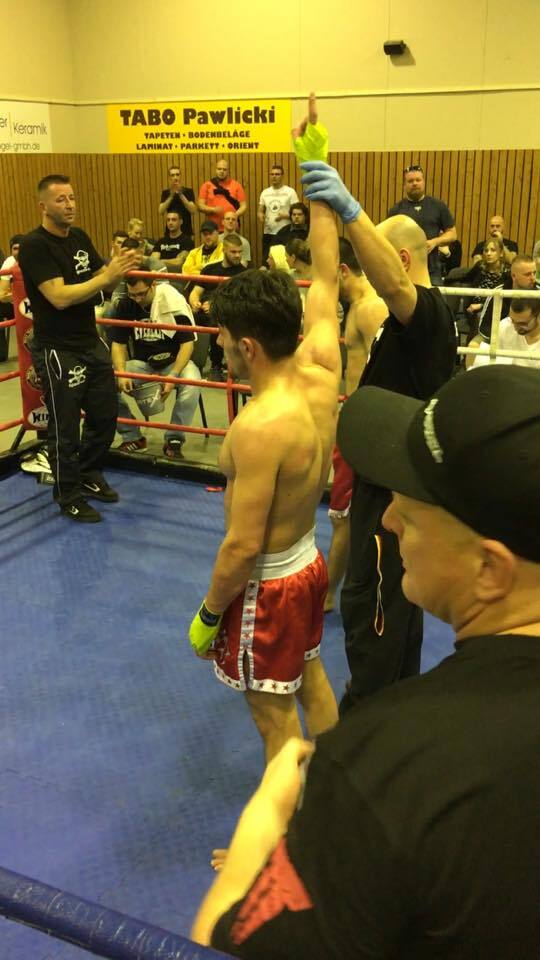 Siegerehrung im Ring: Ein K&auml;mpfer in roten Shorts erh&auml;lt den Sieg, w&auml;hrend Zuschauer und Betreuer zuschauen.