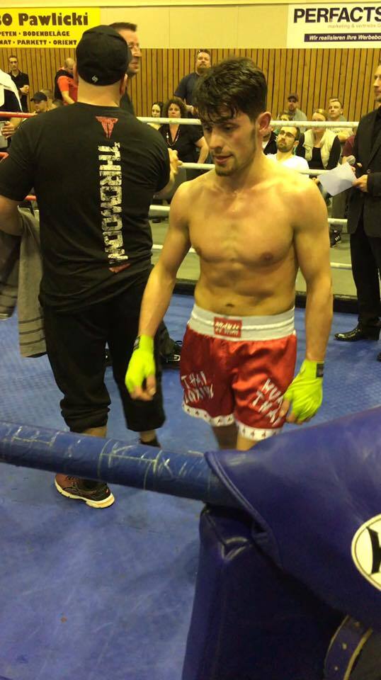 Ein athletischer K&auml;mpfer in roten Shorts und gr&uuml;nen Handschuhen steht in einer Boxarena, umgeben von Zuschauern.