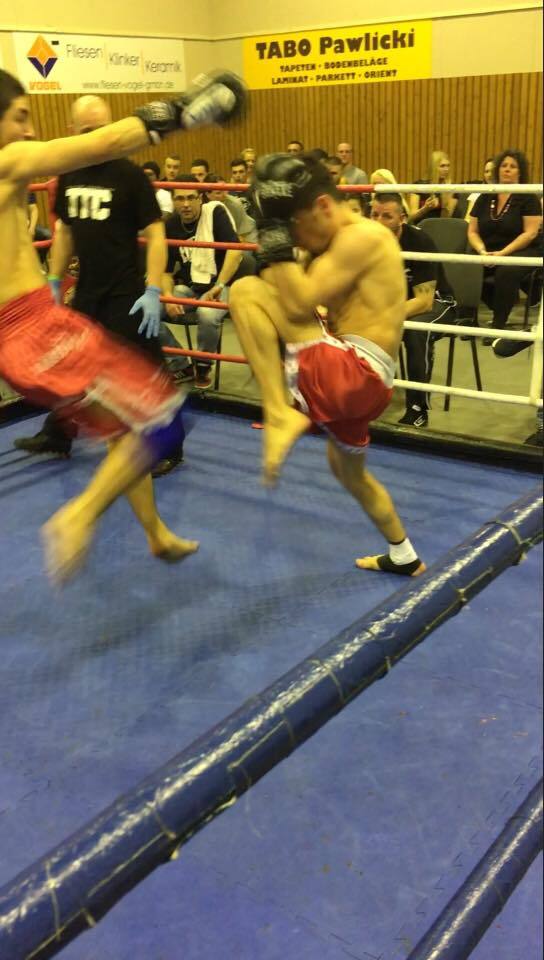 Zwei K&auml;mpfer im Ring, einer f&uuml;hrt einen Kick aus, w&auml;hrend der andere sich abduckt. Zuschauer im Hintergrund.