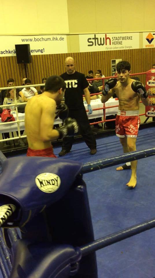 Zwei K&auml;mpfer im Ring, einer tr&auml;gt rote Shorts, der andere schwarze, w&auml;hrend ein Schiedsrichter beobachtet.