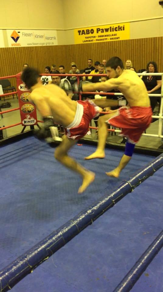 Zwei K&auml;mpfer im Ring f&uuml;hren einen Kickkampf aus, w&auml;hrend Zuschauer auf der Trib&uuml;ne zuschauen.