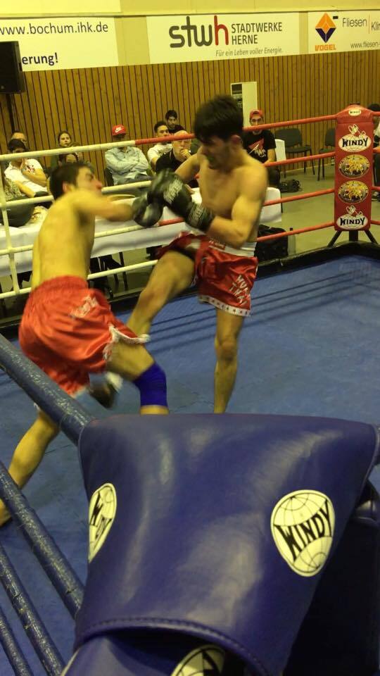 Zwei K&auml;mpfer im Ring, einer mit roter Shorts schl&auml;gt mit einem Fu&szlig;kick auf den anderen ein, Zuschauer im Hintergrund.