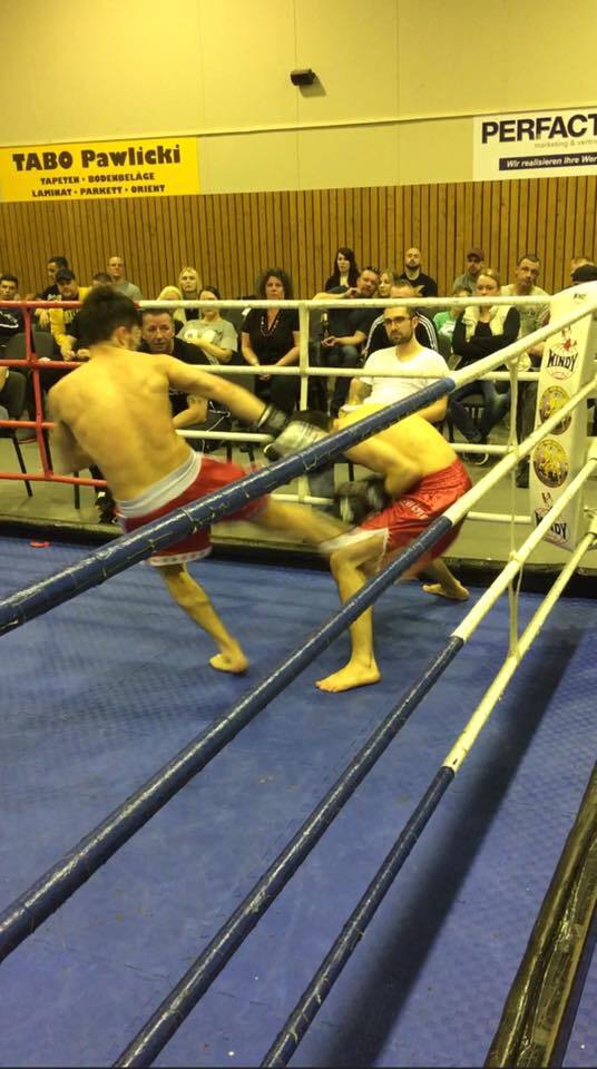 Zwei K&auml;mpfer im Ring w&auml;hrend eines Boxkampfs, ein K&auml;mpfer tritt einen anderen in den Oberschenkel. Zuschauer im Hintergrund.