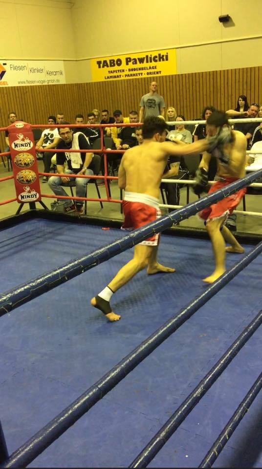 Zwei K&auml;mpfer im Boxring exchange Schl&auml;ge, w&auml;hrend Zuschauer im Hintergrund zuschauen. Rote und blaue Ringe umgeben die K&auml;mpfer.