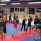Gruppe von Kindern im Training, die mit Boxhandschuhen auf einer blauen Matte vor einem Sandsack &uuml;ben.