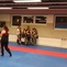 Gruppe von Kindern und Erwachsenen in Trainingskleidung stehen in einem Kampfsportstudio mit blauen und roten Matten.