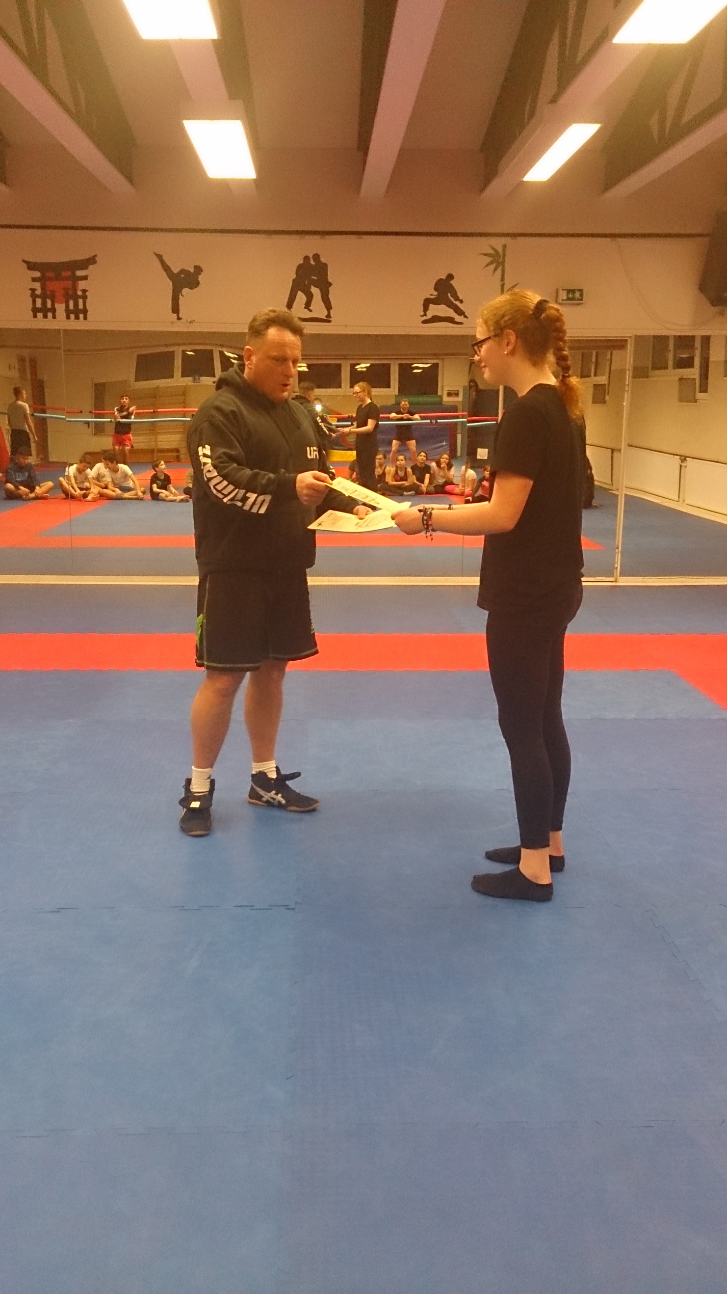 Trainer &uuml;berreicht einer Sportlerin ein Dokument in einer Sporthalle mit Matten und weiteren Teilnehmern im Hintergrund.