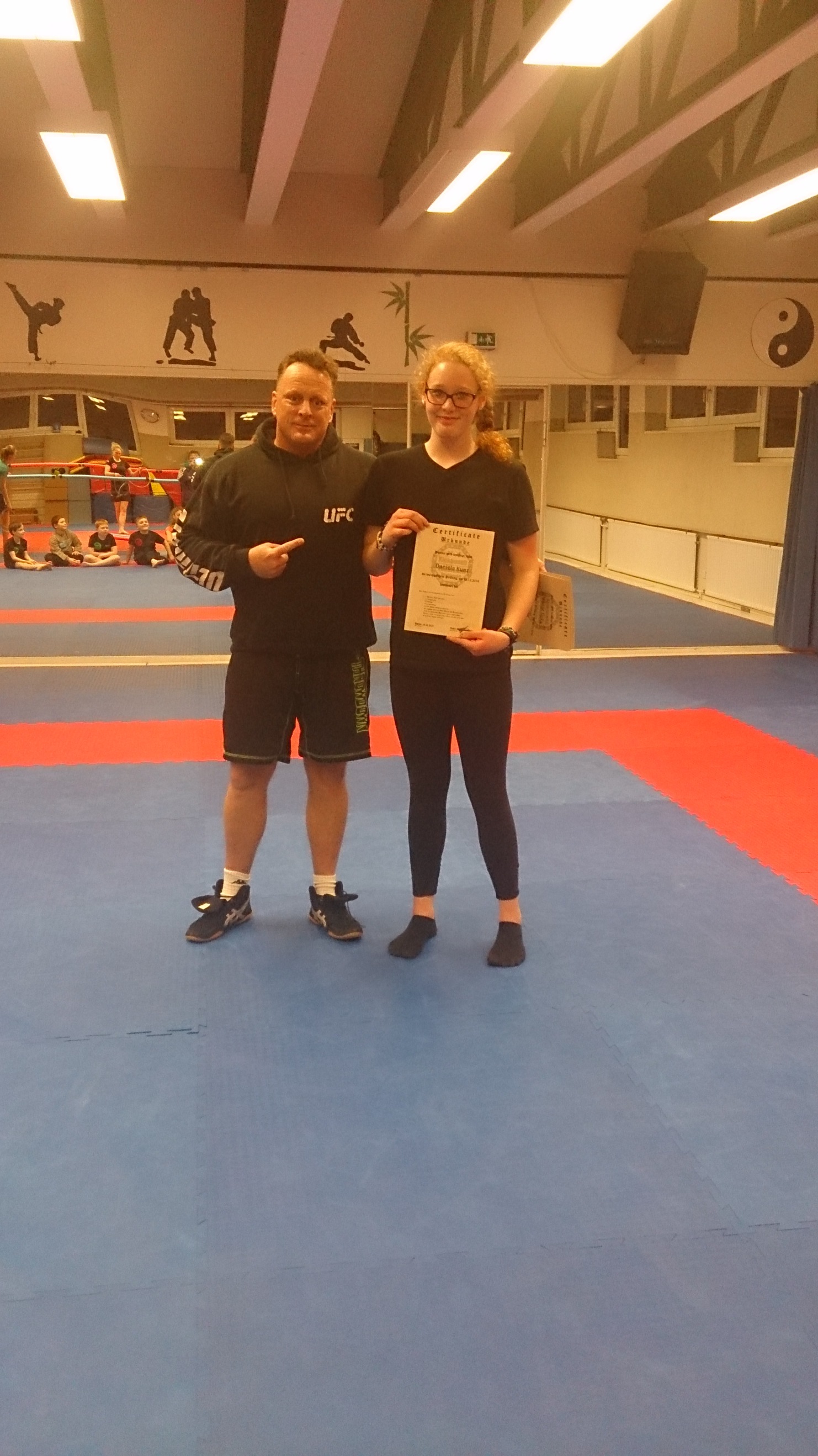 Ein Trainer &uuml;berreicht einer jungen Sportlerin eine Urkunde in einem Kampfsportstudio mit Matten und Dekoration.