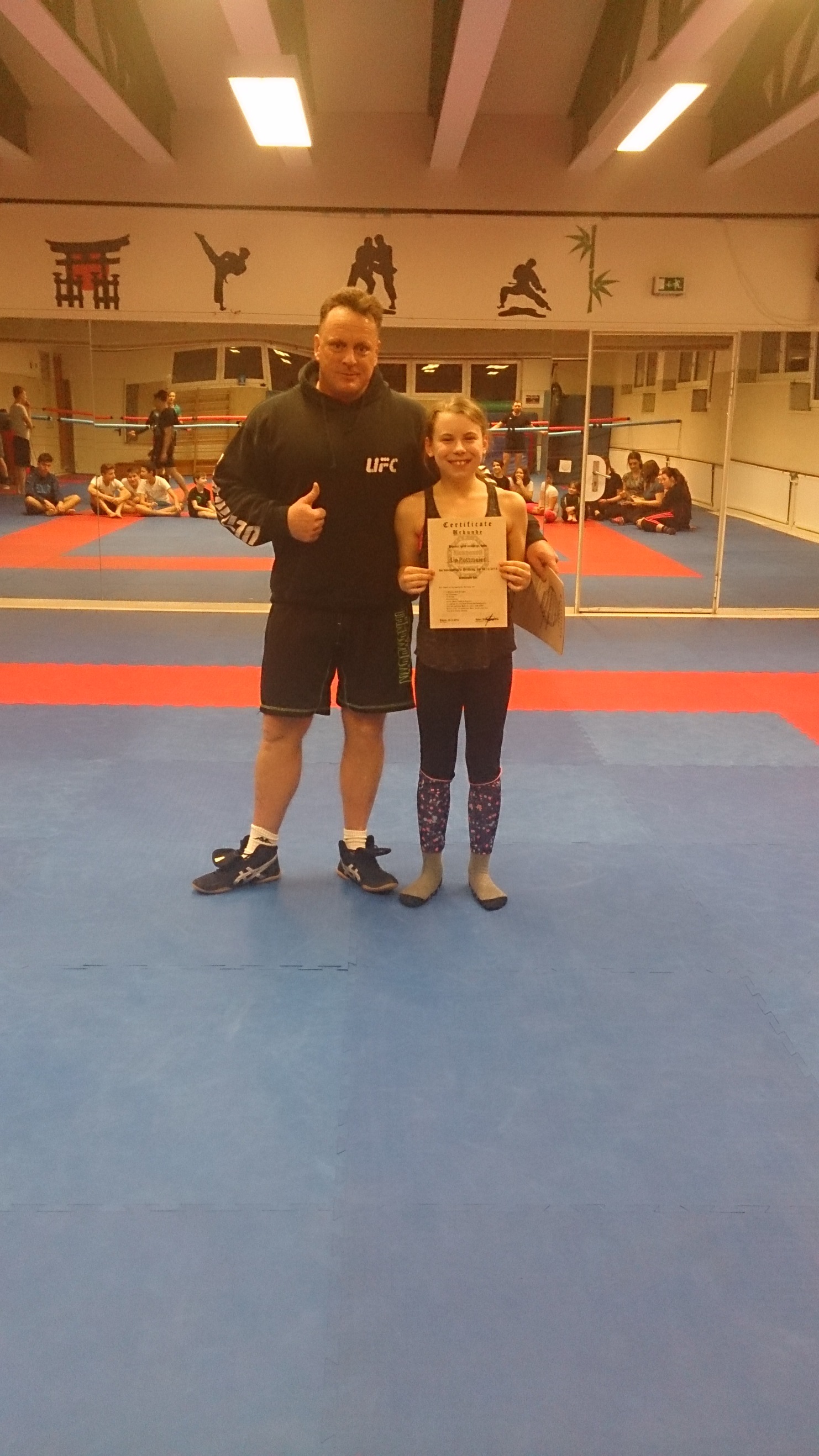 Sportler und junges M&auml;dchen stehen auf einer Trainingsmatte, das M&auml;dchen h&auml;lt eine Urkunde und l&auml;chelt stolz.