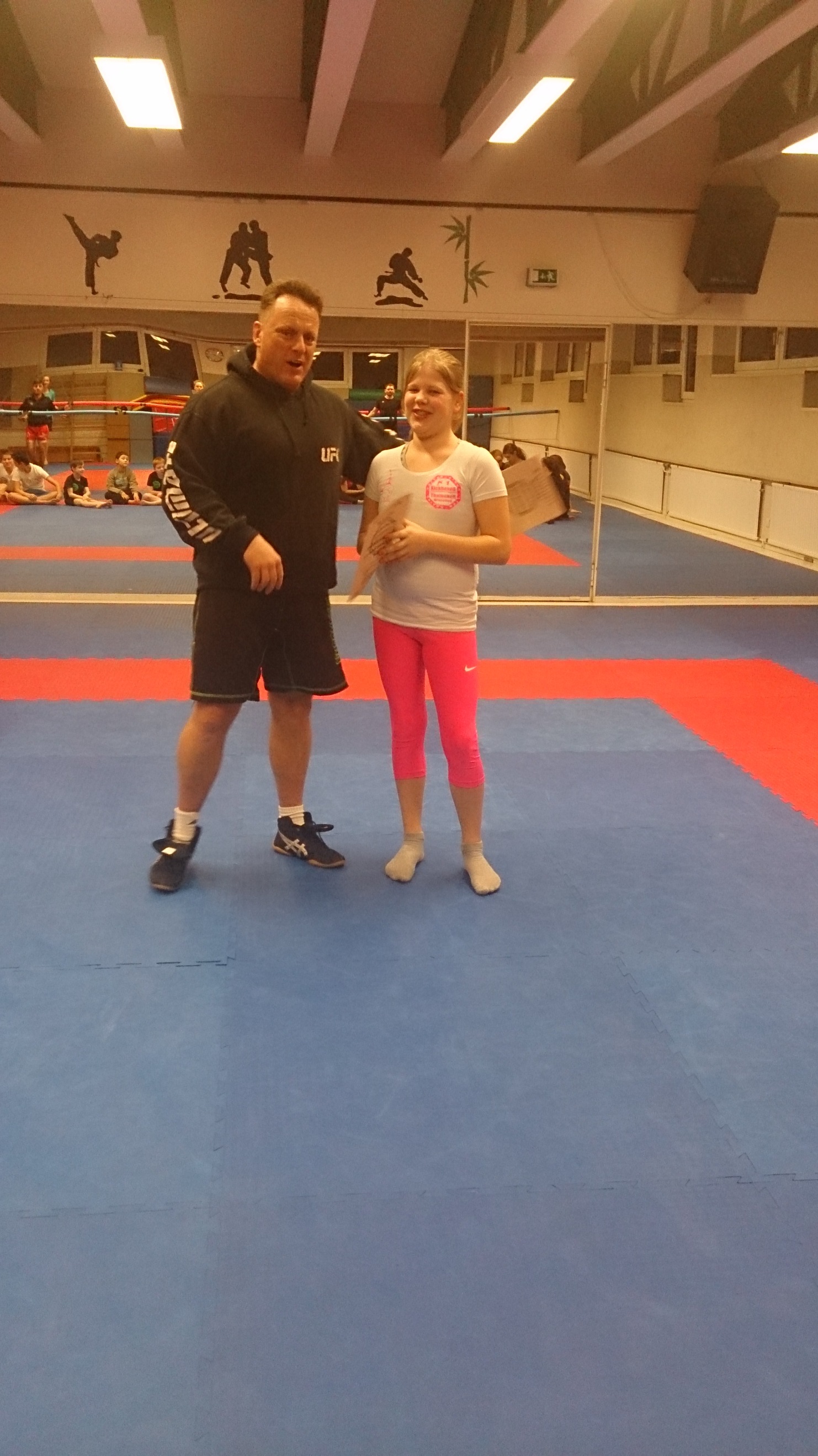 Zwei Personen stehen auf einer blauen Sportmatte in einem Trainingsraum, w&auml;hrend im Hintergrund andere Aktivit&auml;t sichtbar ist.