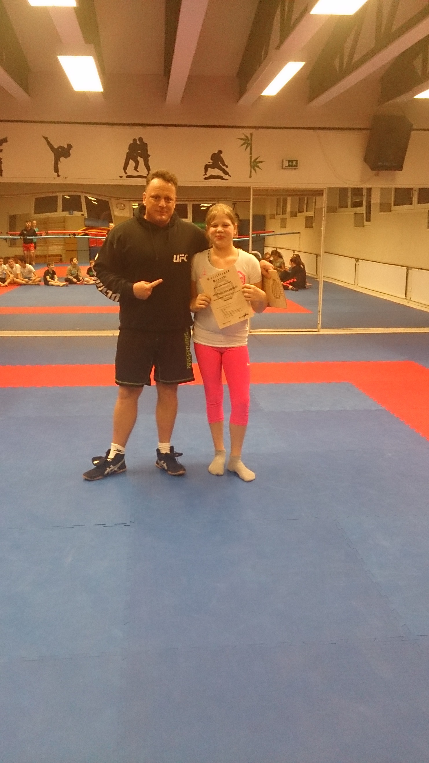 Junge Sportlerin h&auml;lt Urkunde, steht neben Trainer in einer Sporthalle mit Teppichboden. Hintergrund zeigt weitere Kinder beim Training.