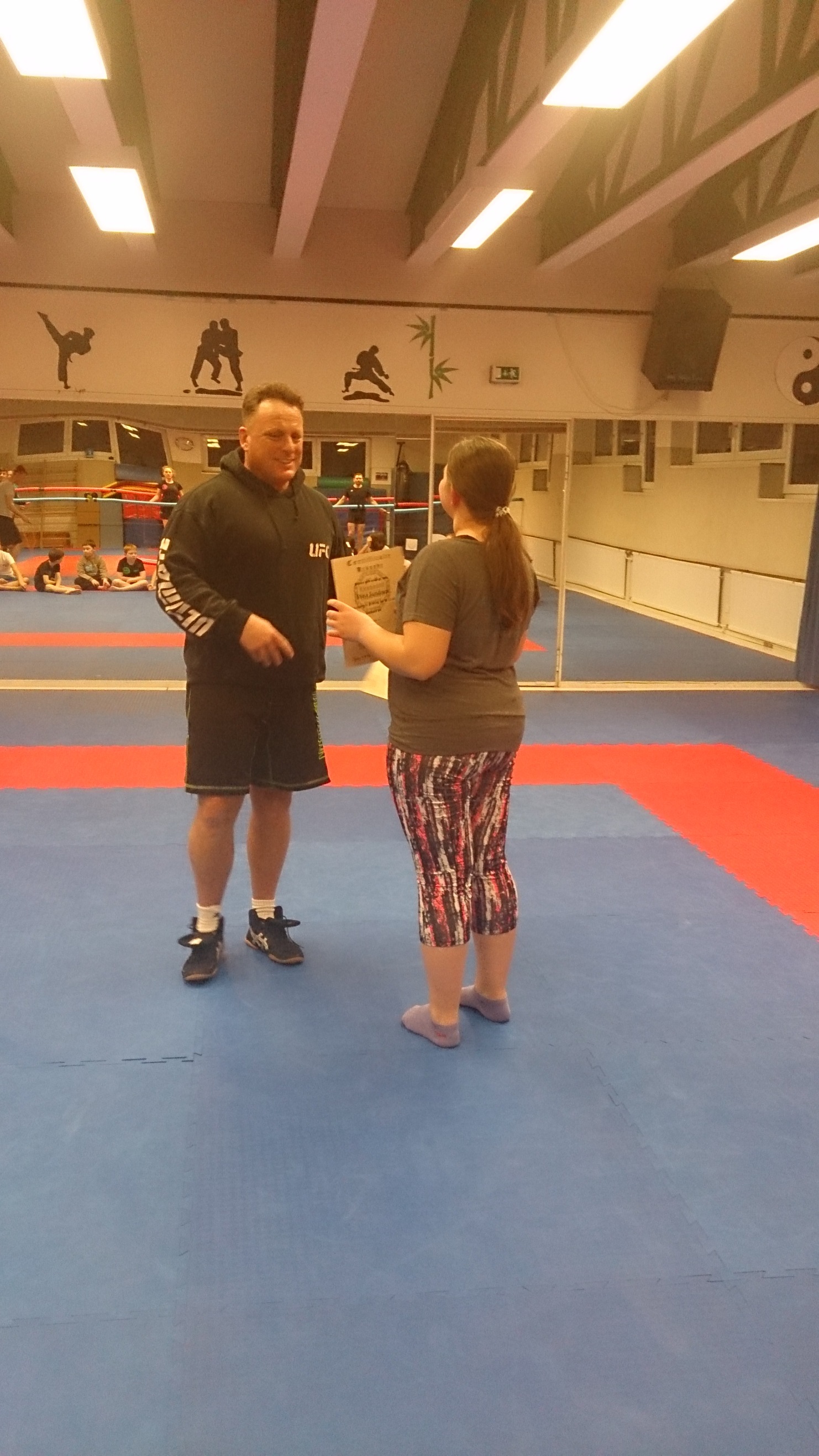 Zwei Sportler in Trainingskleidung stehen auf einer blauen Matte in einem Kampfsportstudio.