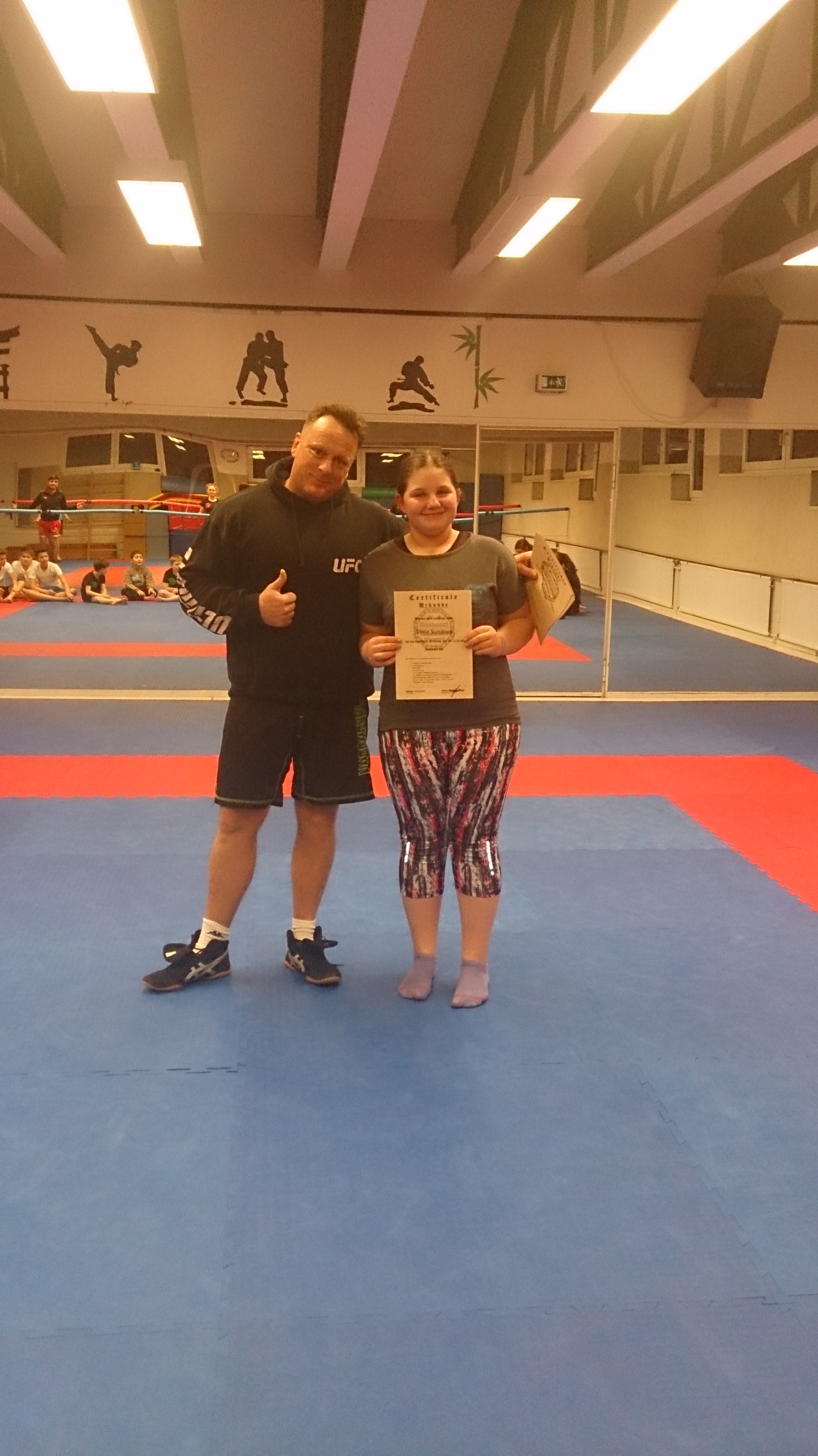 Zwei Personen stehen auf einer Trainingsmatte, eine h&auml;lt ein Zertifikat, der andere zeigt Daumen hoch.