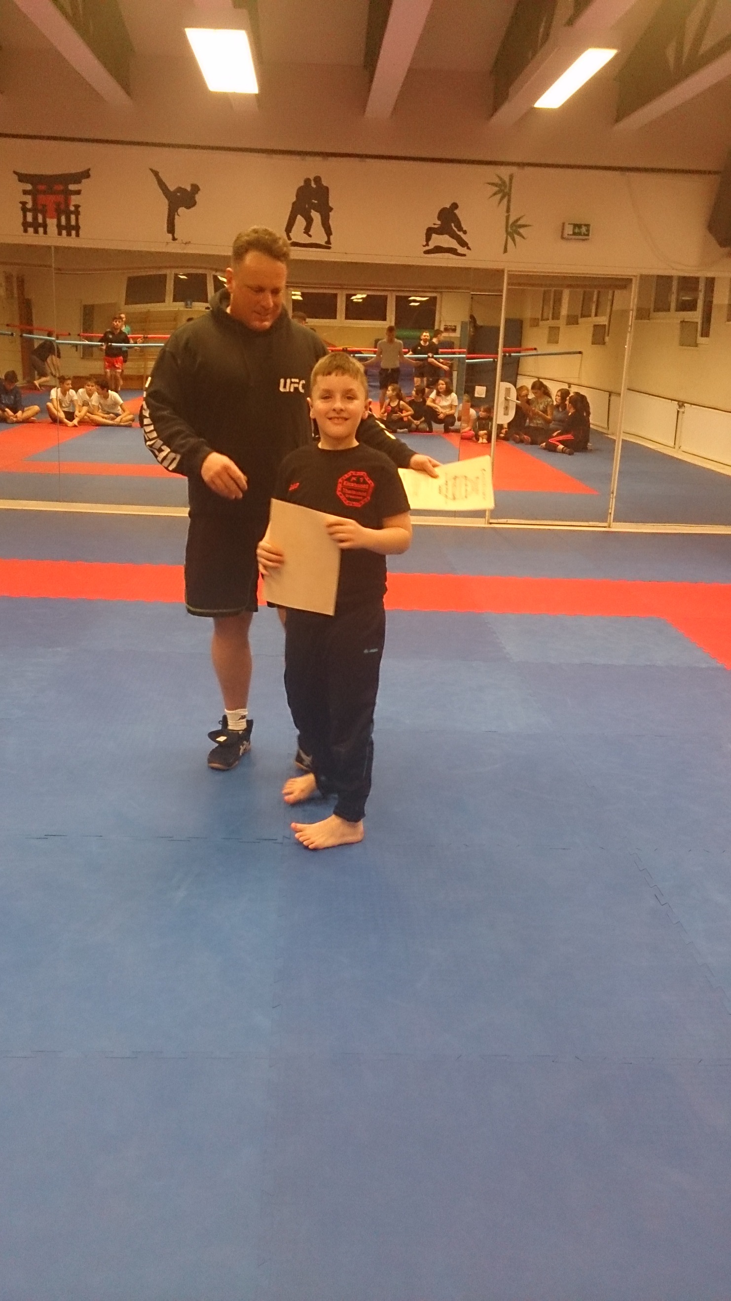 Ein Trainer &uuml;berreicht einem Jungen eine Urkunde in einem Kampfsportzentrum mit blauen Matten.