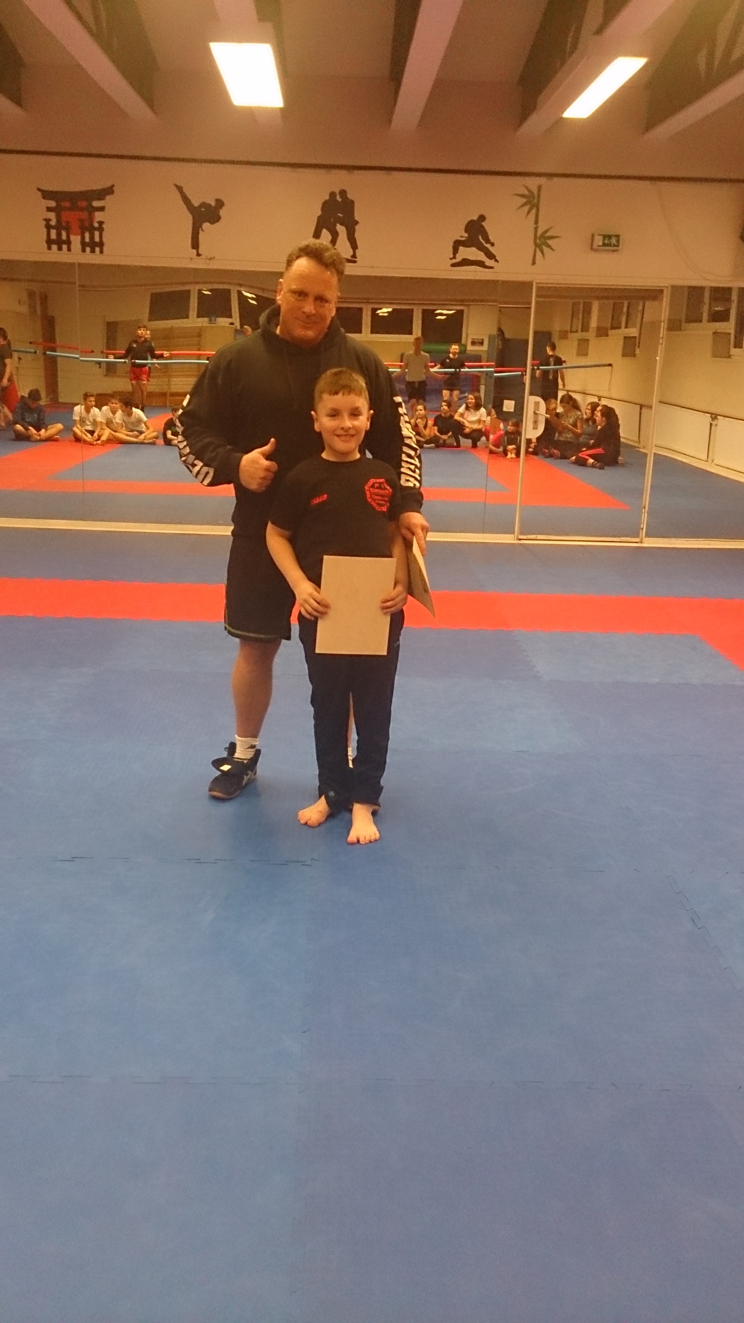 Trainer mit Daumen hoch steht neben einem Jungen, der ein Zertifikat in der Hand h&auml;lt, auf einer Kampfsportmatte.