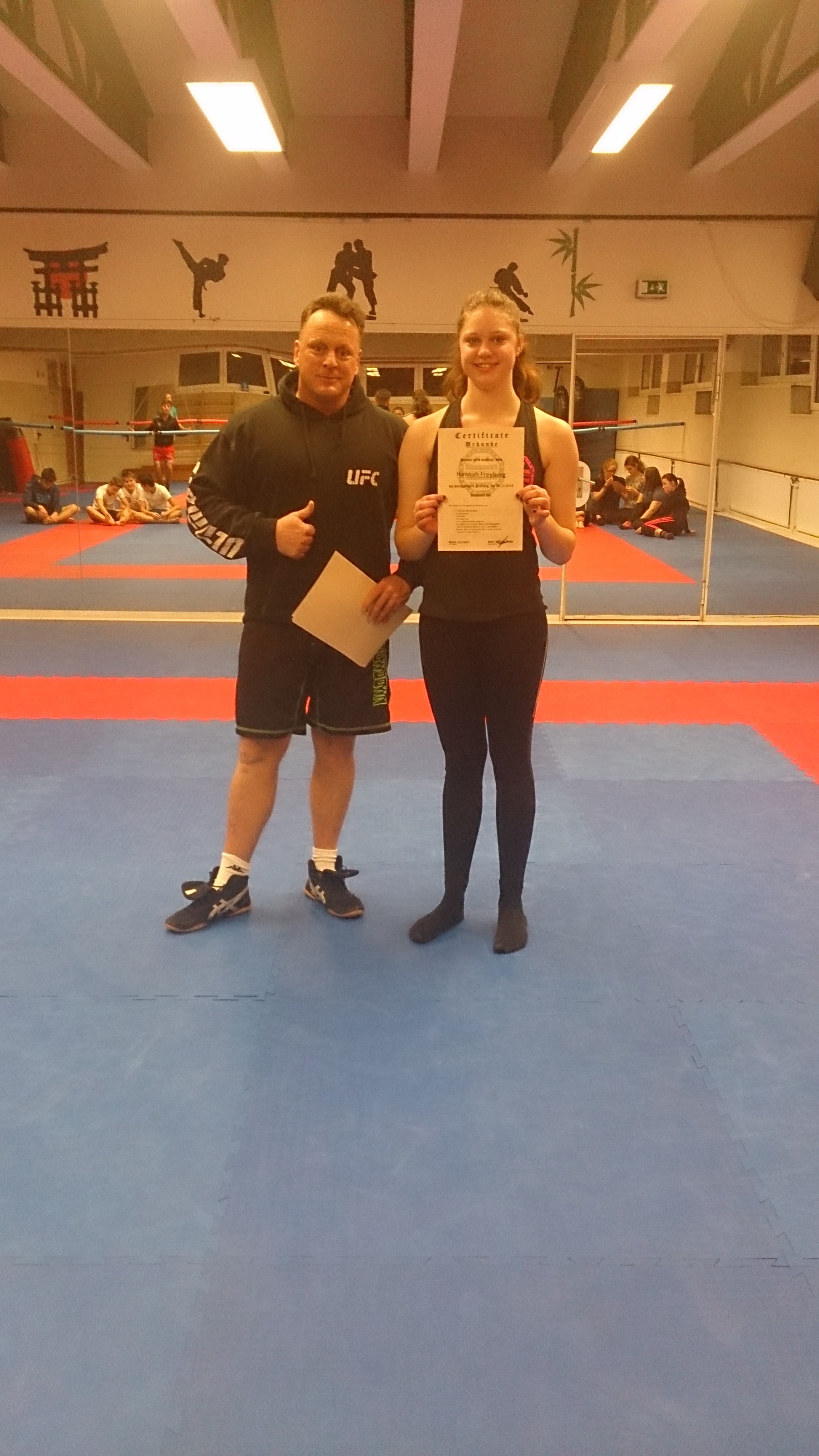 Mann und Frau stehen auf einer Kampfsportmatte; die Frau h&auml;lt ein Zertifikat, der Mann zeigt Daumen hoch.