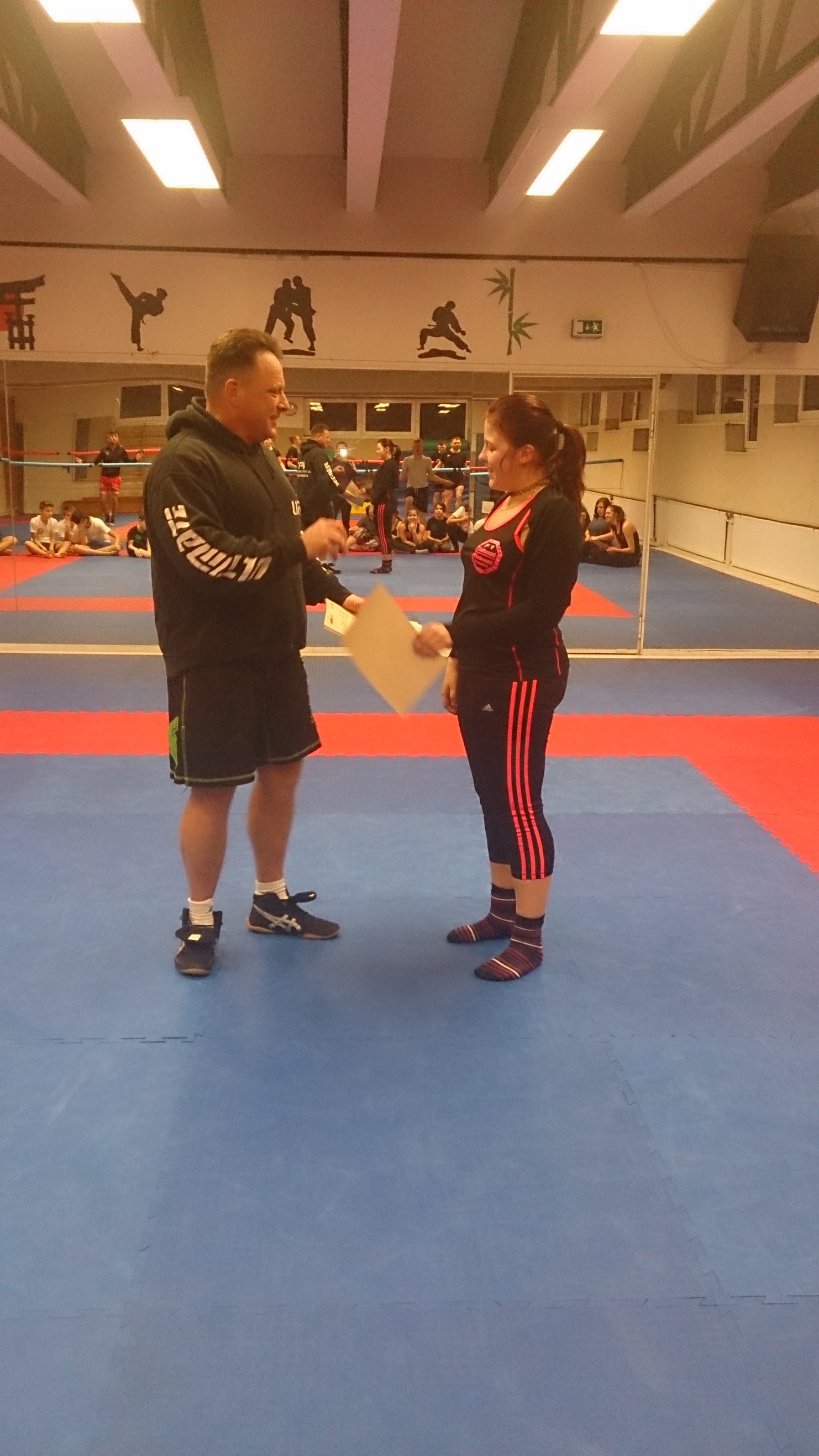 Trainer &uuml;berreicht einer Sportlerin eine Urkunde in einer Kampfsporthalle mit Spiegeln und Zuschauern im Hintergrund.