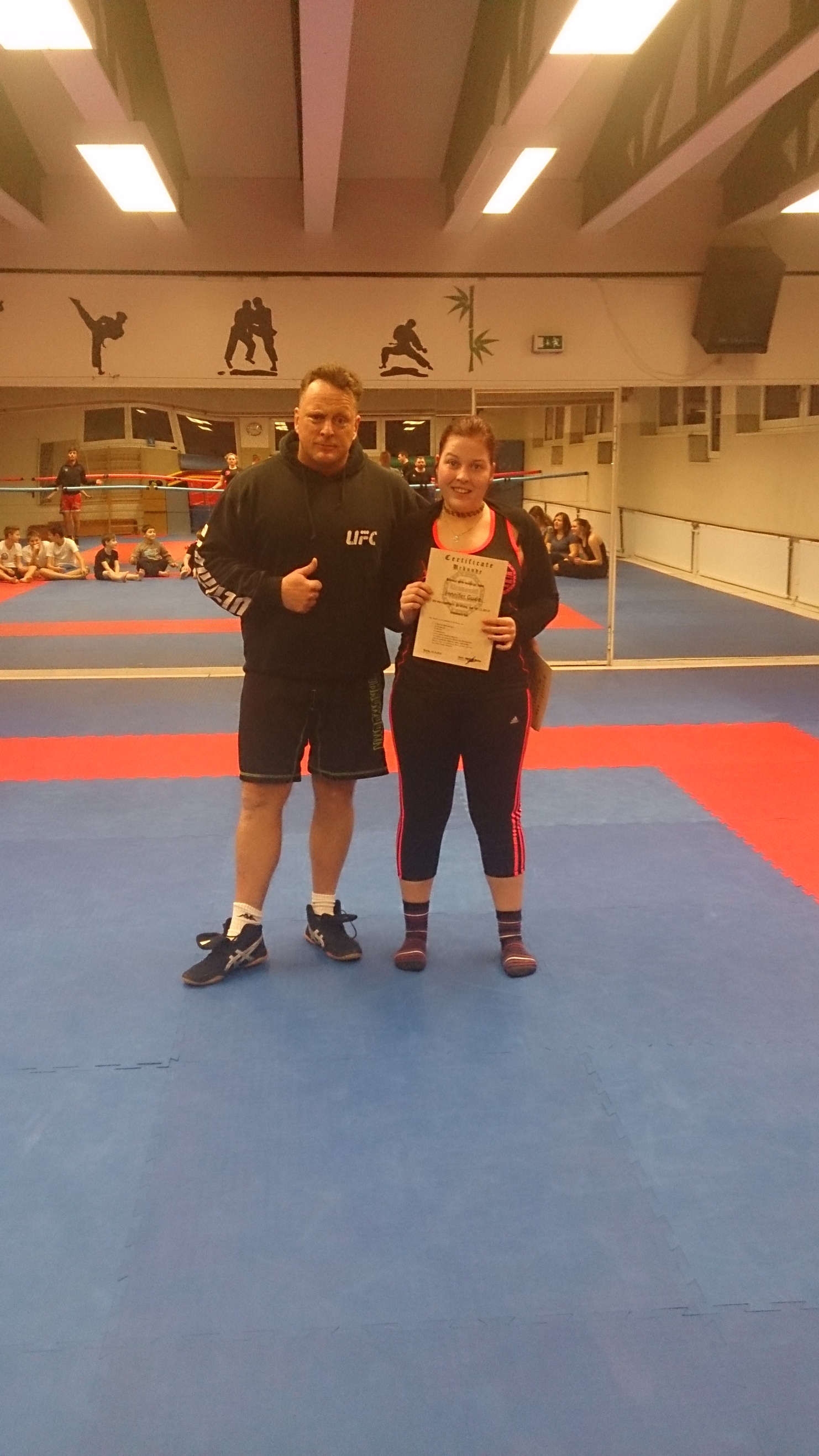 Ein Mann und eine Frau stehen in einer Sporthalle, die Frau h&auml;lt ein Diplom, beide l&auml;cheln freundlich.