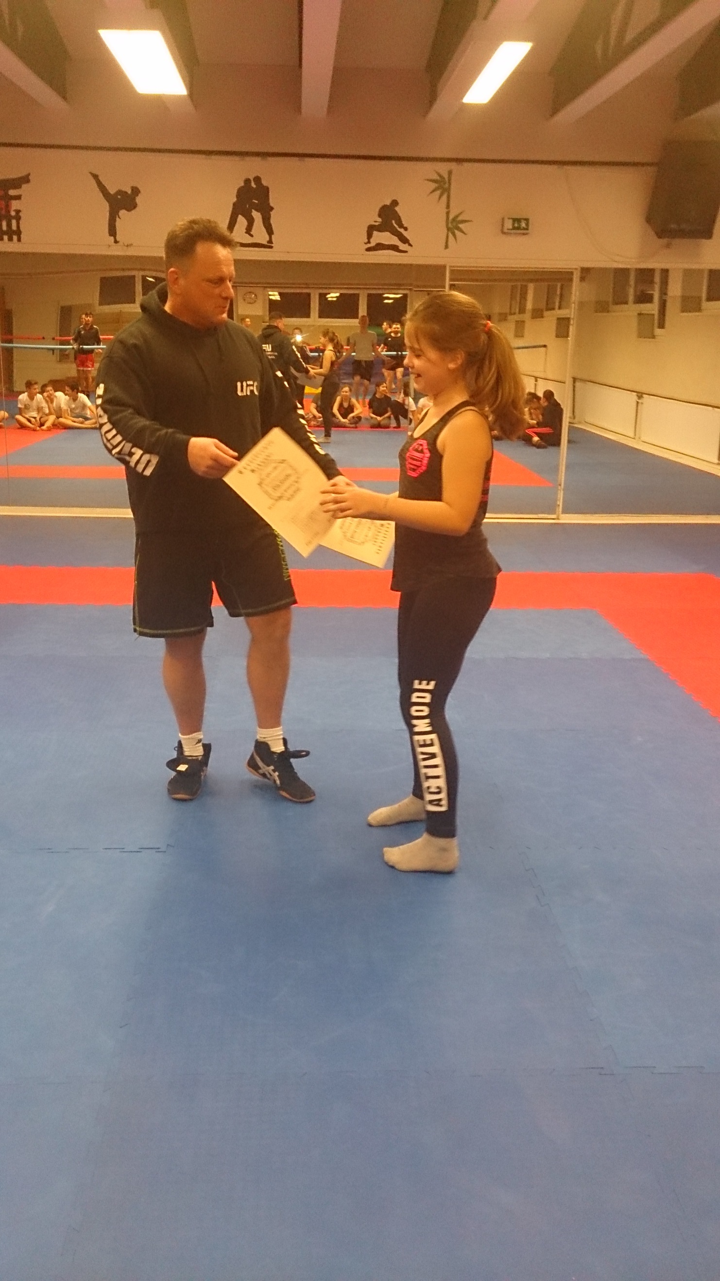 Ein Trainer &uuml;berreicht einer jungen Sportlerin eine Urkunde in einem Kampfsportstudio.