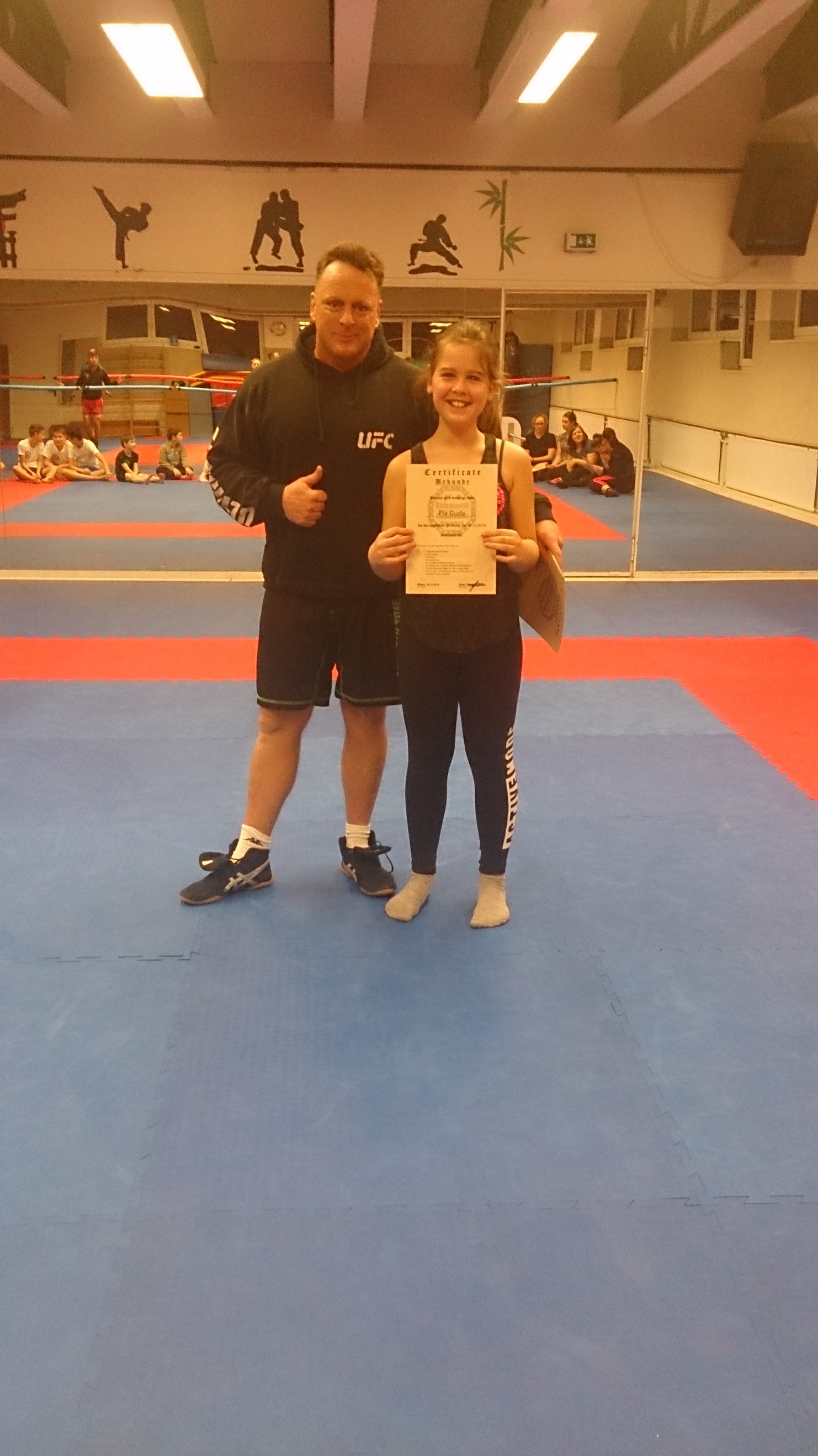 Trainer und junge K&auml;mpferin halten ein Zertifikat auf einer blauen und roten Kampfsportmatte in einem Trainingsraum.