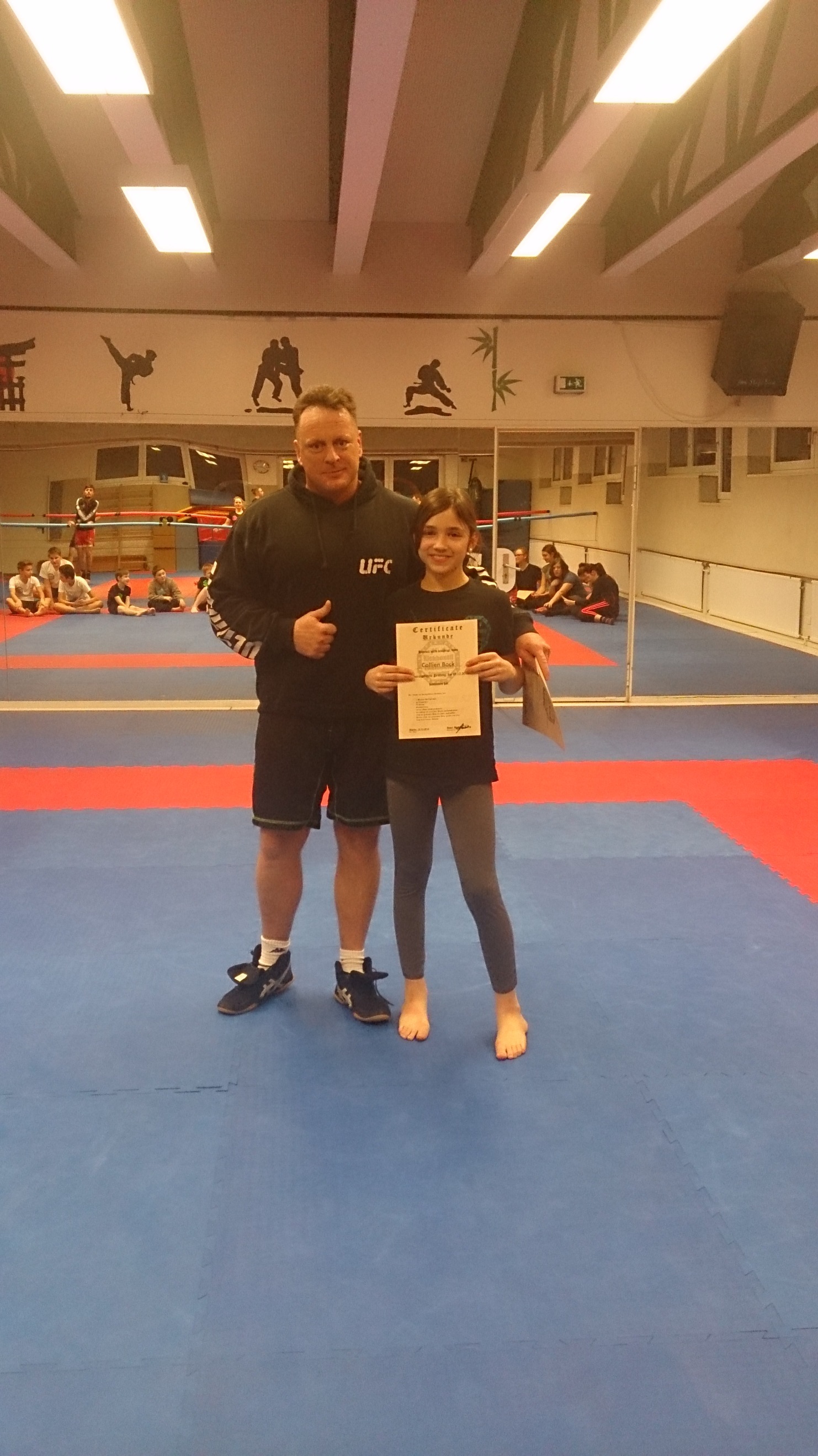 Ein Trainer und ein M&auml;dchen halten stolz ein Zertifikat in einem Kampfsportstudio mit blauen Matten.