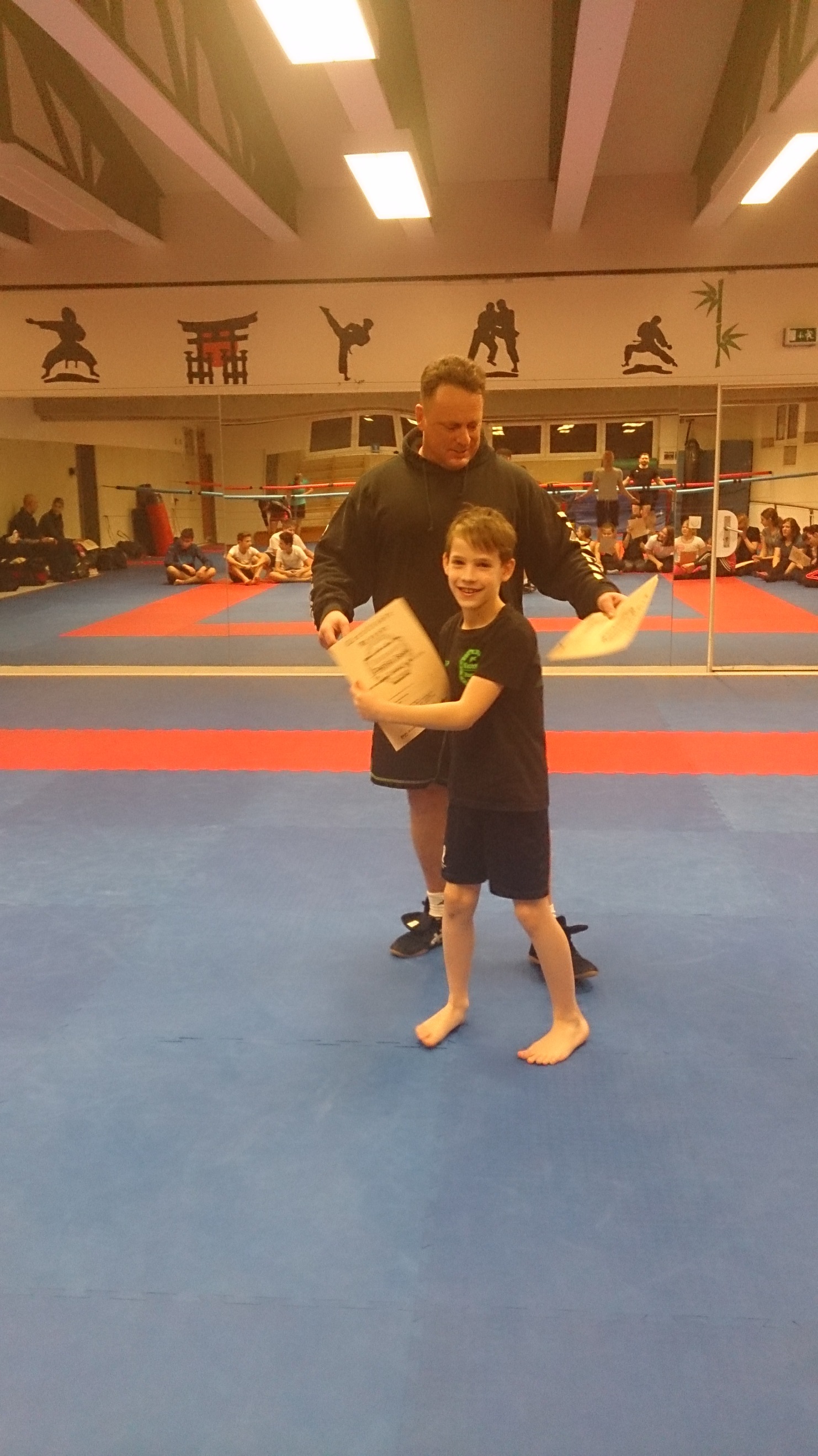 Ein Trainer &uuml;berreicht einem Jungen ein Dokument in einer Sporthalle mit Trainingsmatten und Zuschauern im Hintergrund.