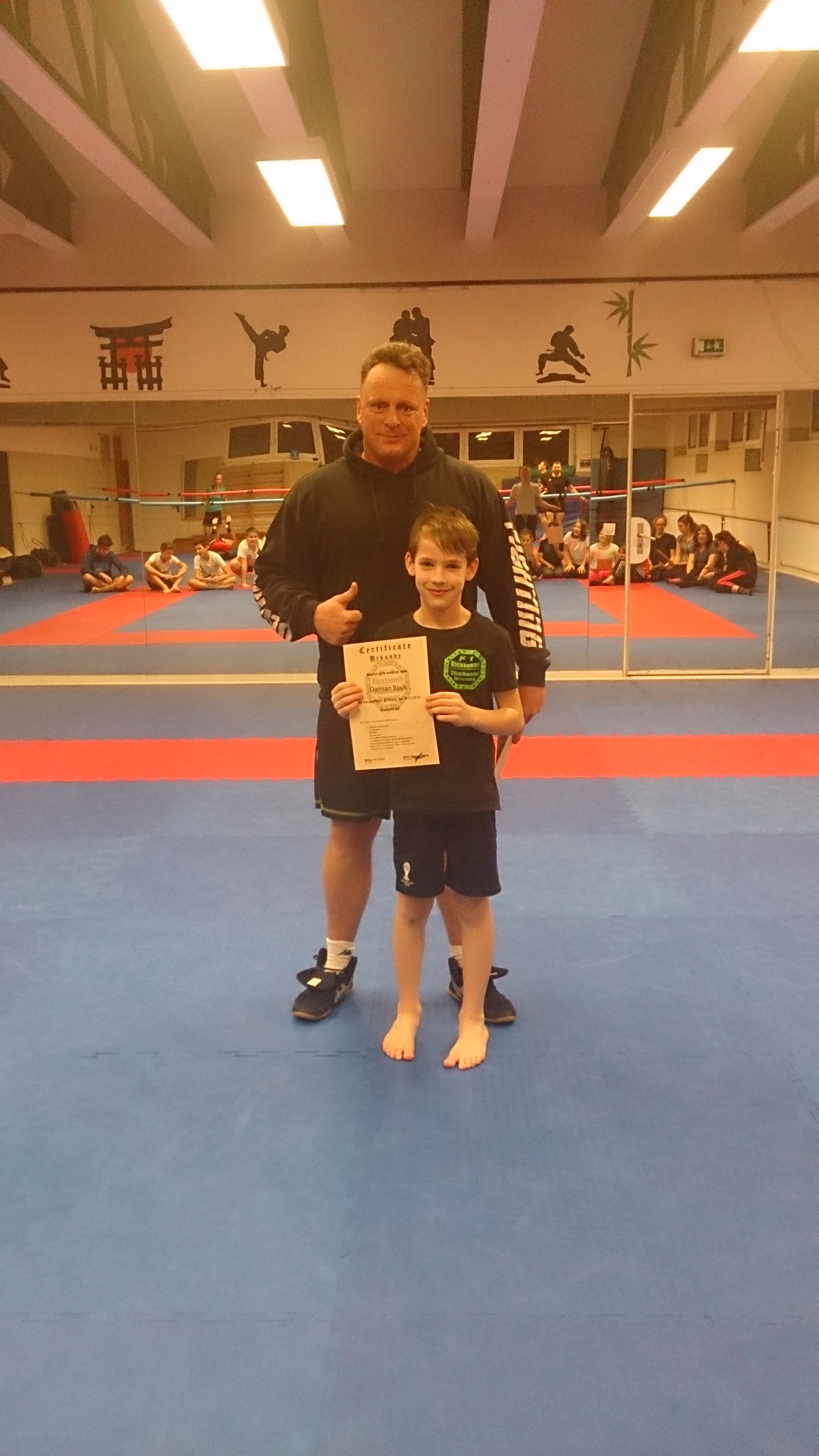 Trainer steht mit einem Jungen, der ein Zertifikat in der Hand h&auml;lt, auf einer blauen Kampfsportmatte.