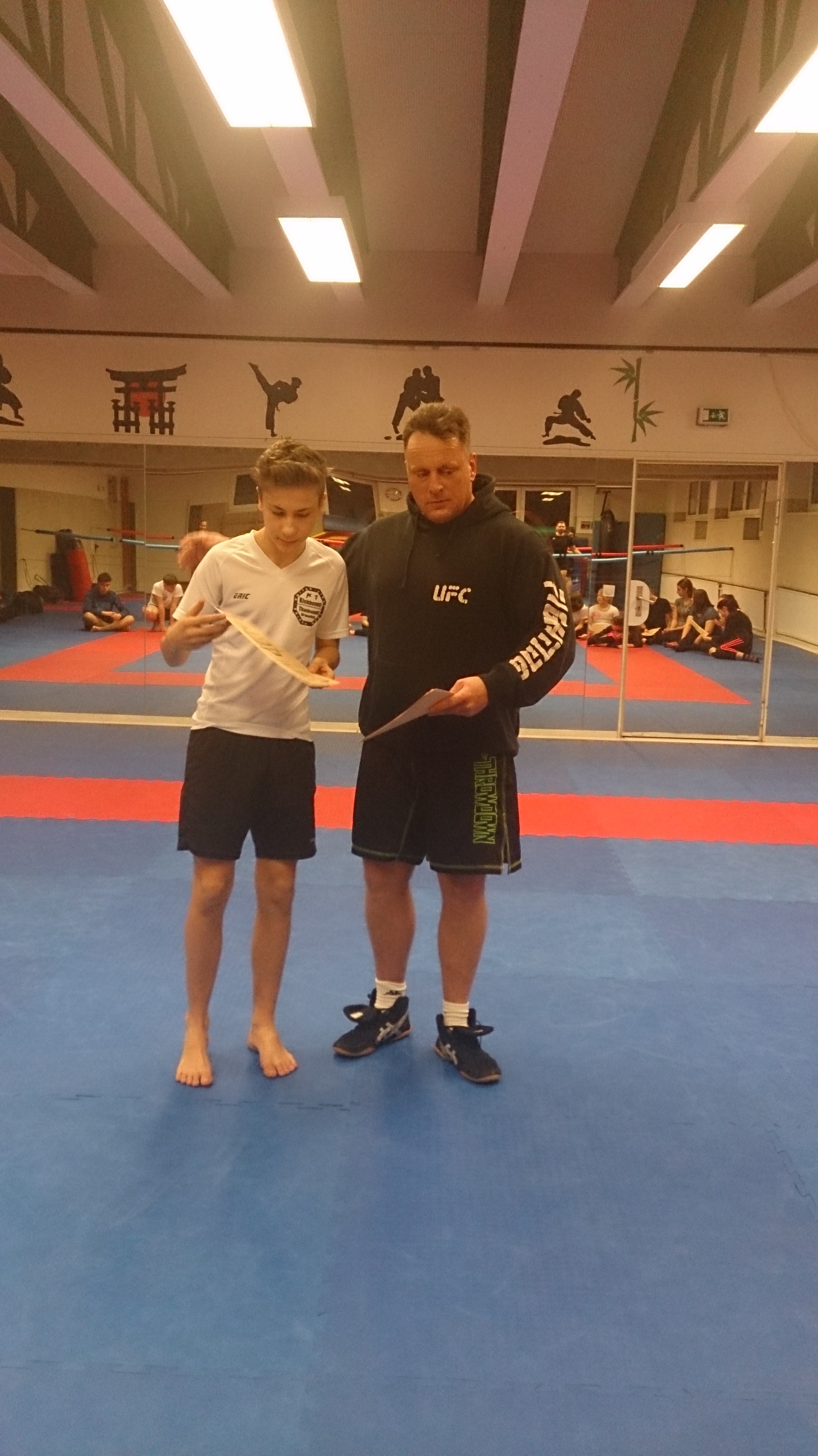 Zwei M&auml;nner stehen in einem Trainingsraum, einer zeigt einem anderen eine Technik mit einem Trainingstr&auml;ger.