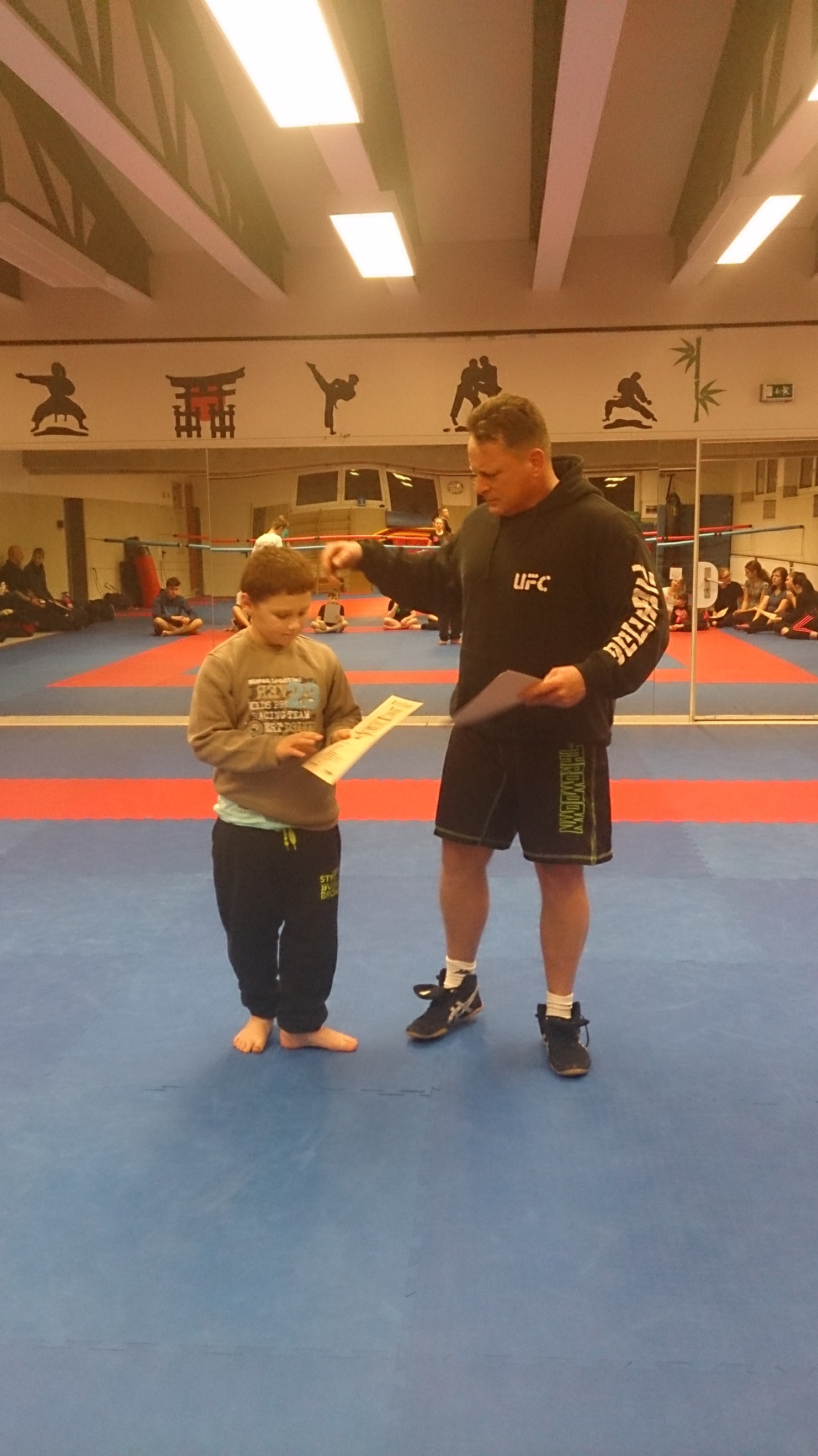 Trainer erkl&auml;rt einem Kindes die Anweisungen auf einem Blatt im Kampfsport-Trainingsraum mit blauer Matte.