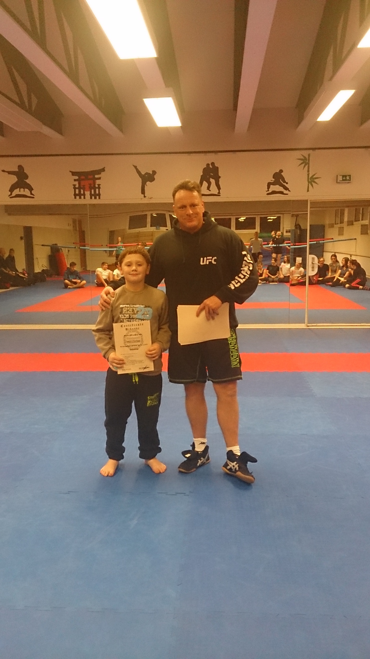 Junge und Trainer posieren auf einer Kampfsportmatte, der Junge h&auml;lt eine Urkunde in der Hand.