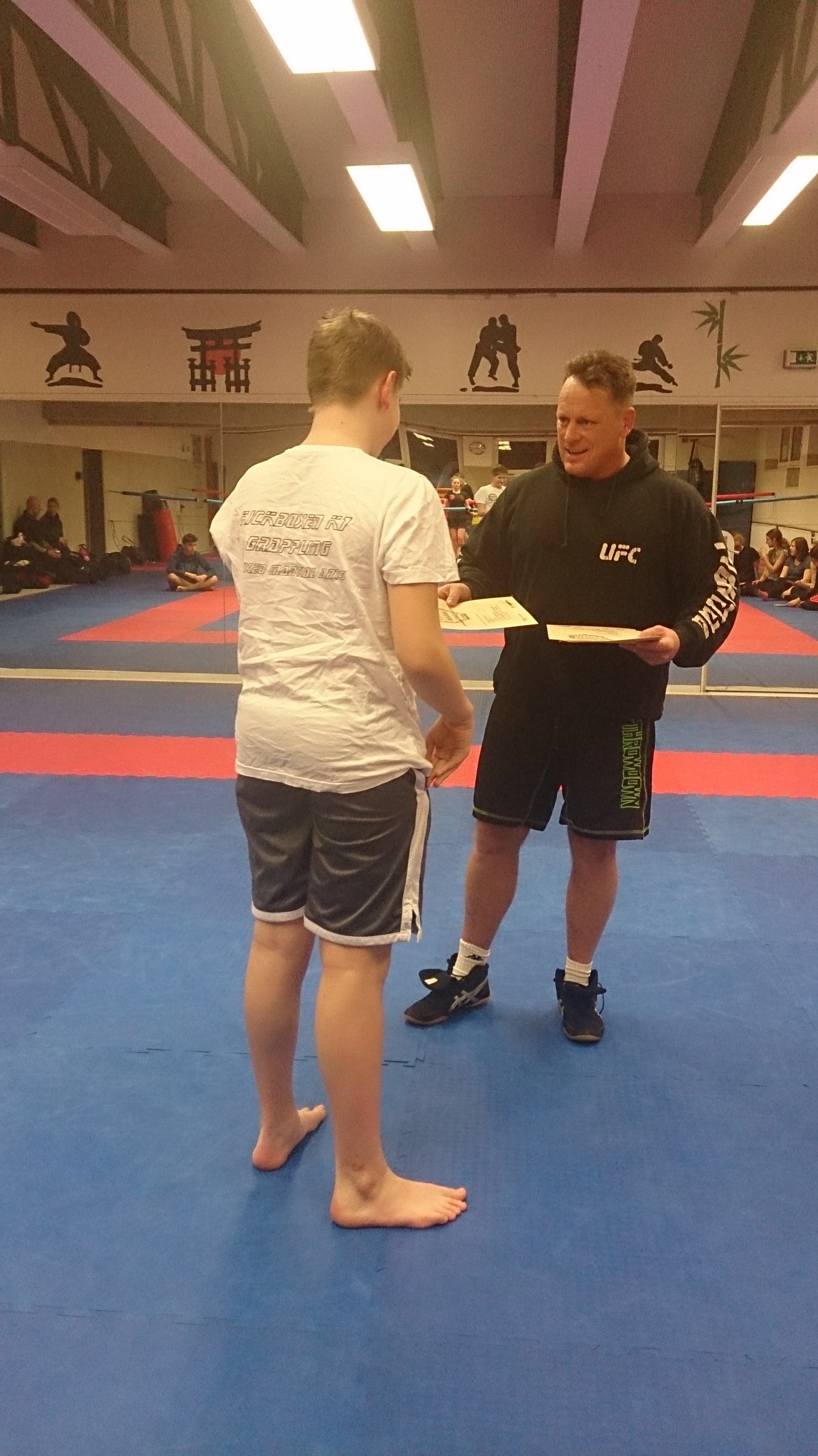 Zwei Personen in Sportkleidung stehen auf einer blauen Matte, ein Mann &uuml;berreicht einem Jungen ein Dokument.