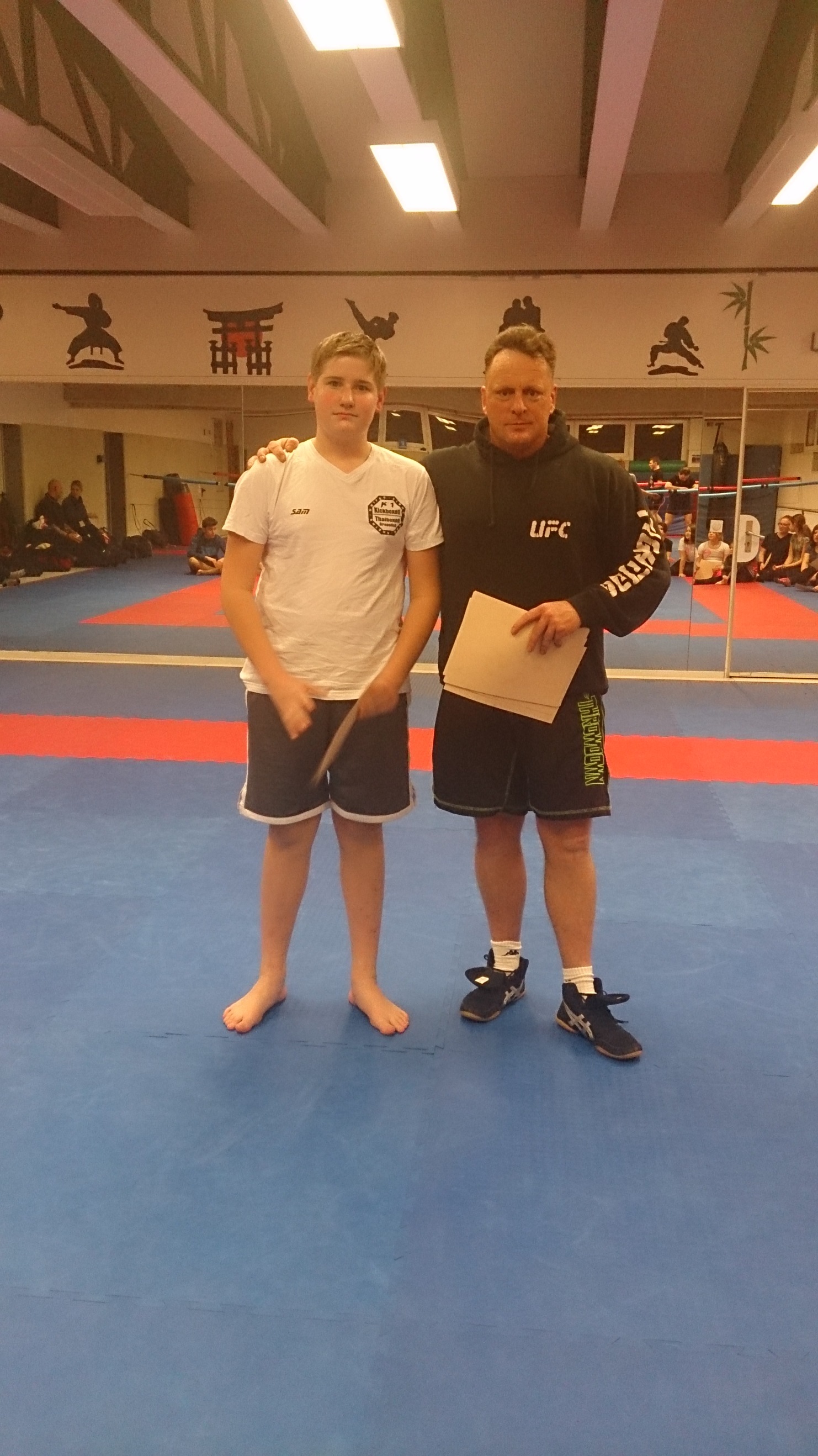 Zwei M&auml;nner stehen auf einer blauen Trainingsmatte. Einer tr&auml;gt ein UFC-Hoodie, der andere ein T-Shirt und Shorts.