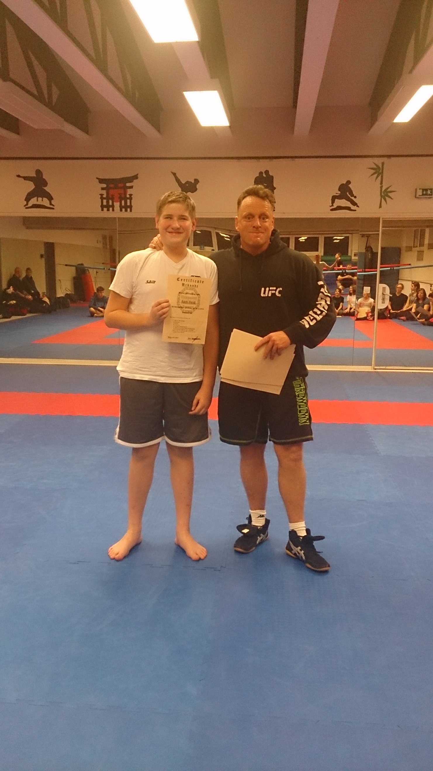 Zwei Personen stehen auf einer Sportmatte. Der Junge h&auml;lt eine Urkunde, der Trainer hat einen Ordner in der anderen Hand.