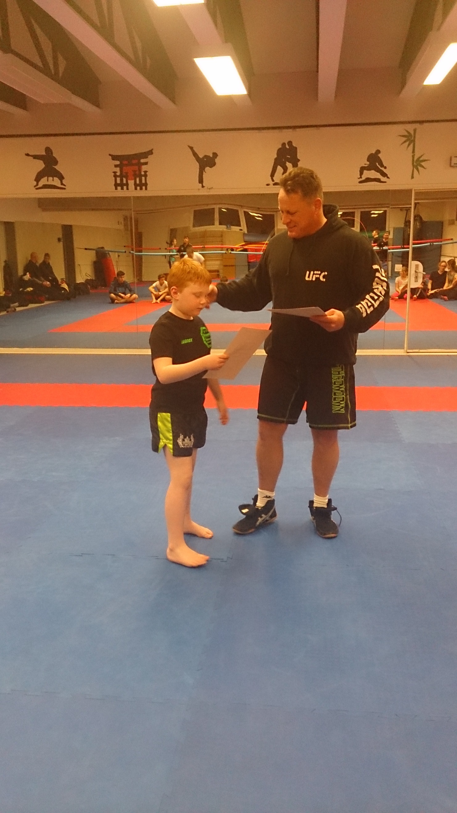 Trainer spricht mit einem Kindesportler auf einer blauen Trainingsfl&auml;che, beide halten Dokumente in der Hand.